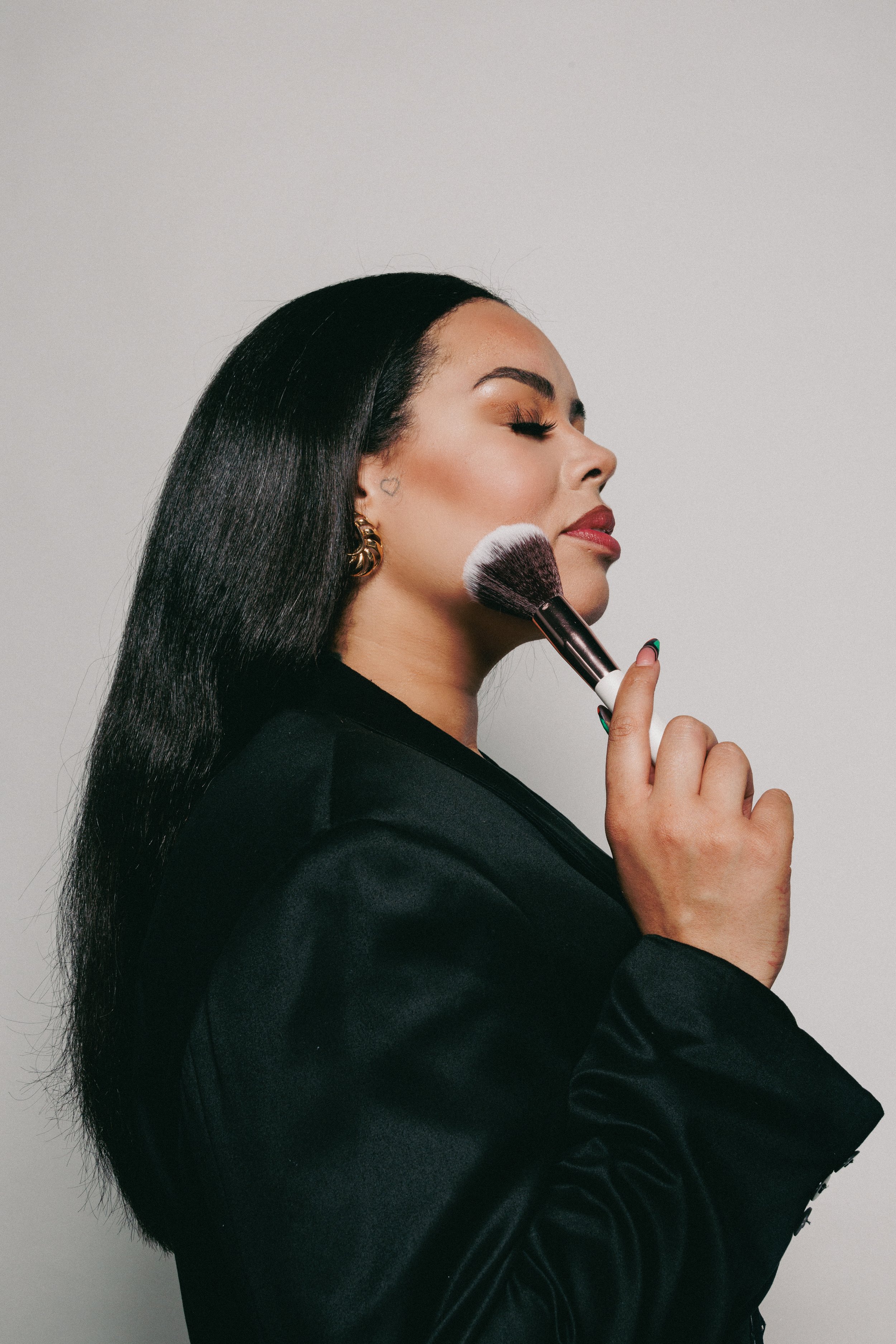 A woman with long black hair, closed eyes, wearing gold earrings, holding a makeup brush to her face against a plain gray background.