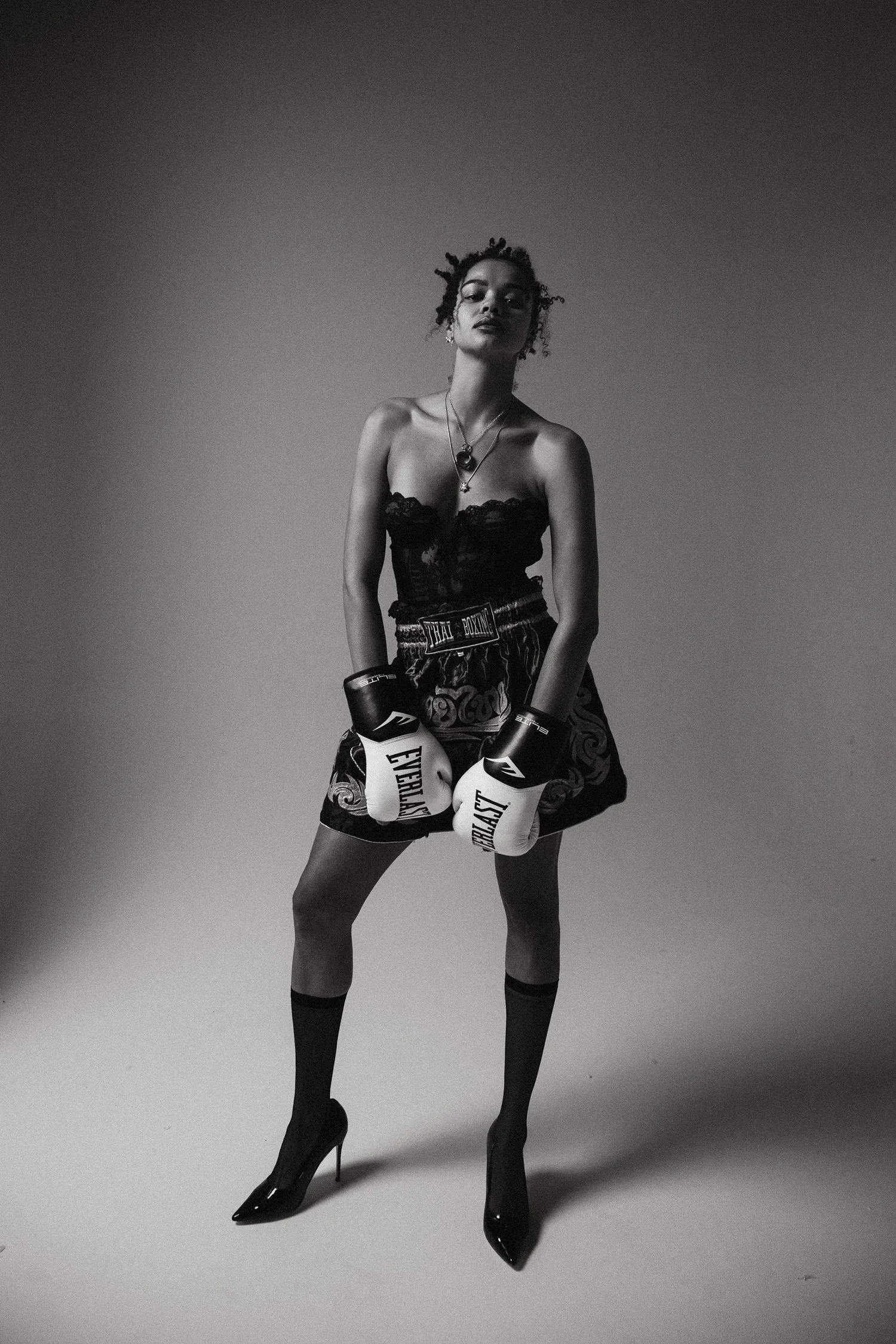 A woman standing confidently in high heels, boxing gloves, and boxing shorts, with jewelry and a confident expression.