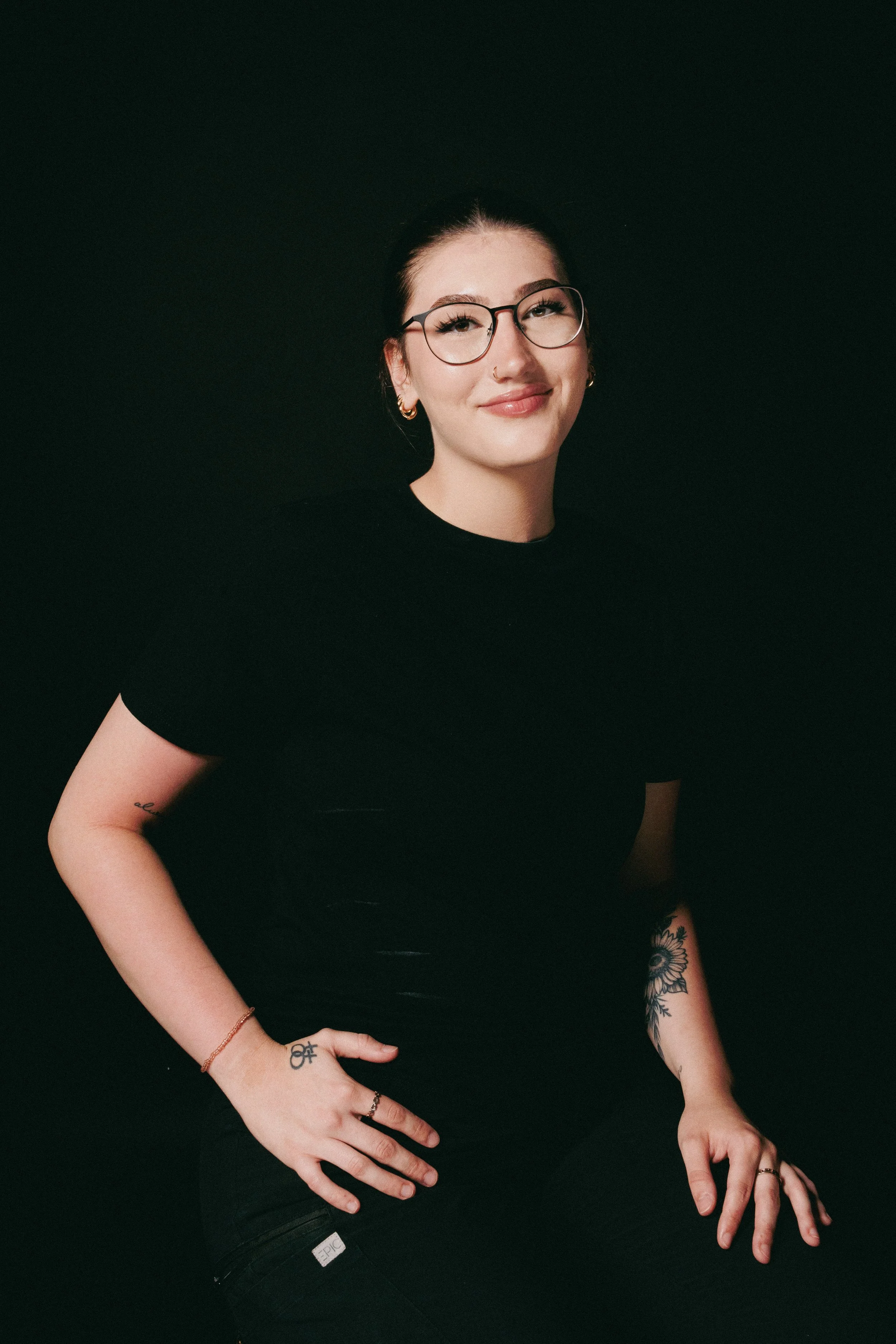 A woman with glasses and tattoos wearing a black shirt, smiling, against a black background.