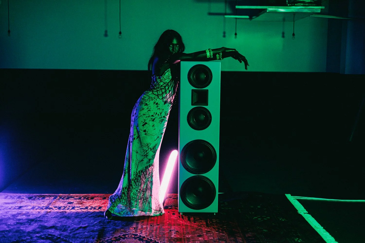 A woman in a colorful patterned dress leaning on a large speaker in a dimly lit room with neon and colored lighting.