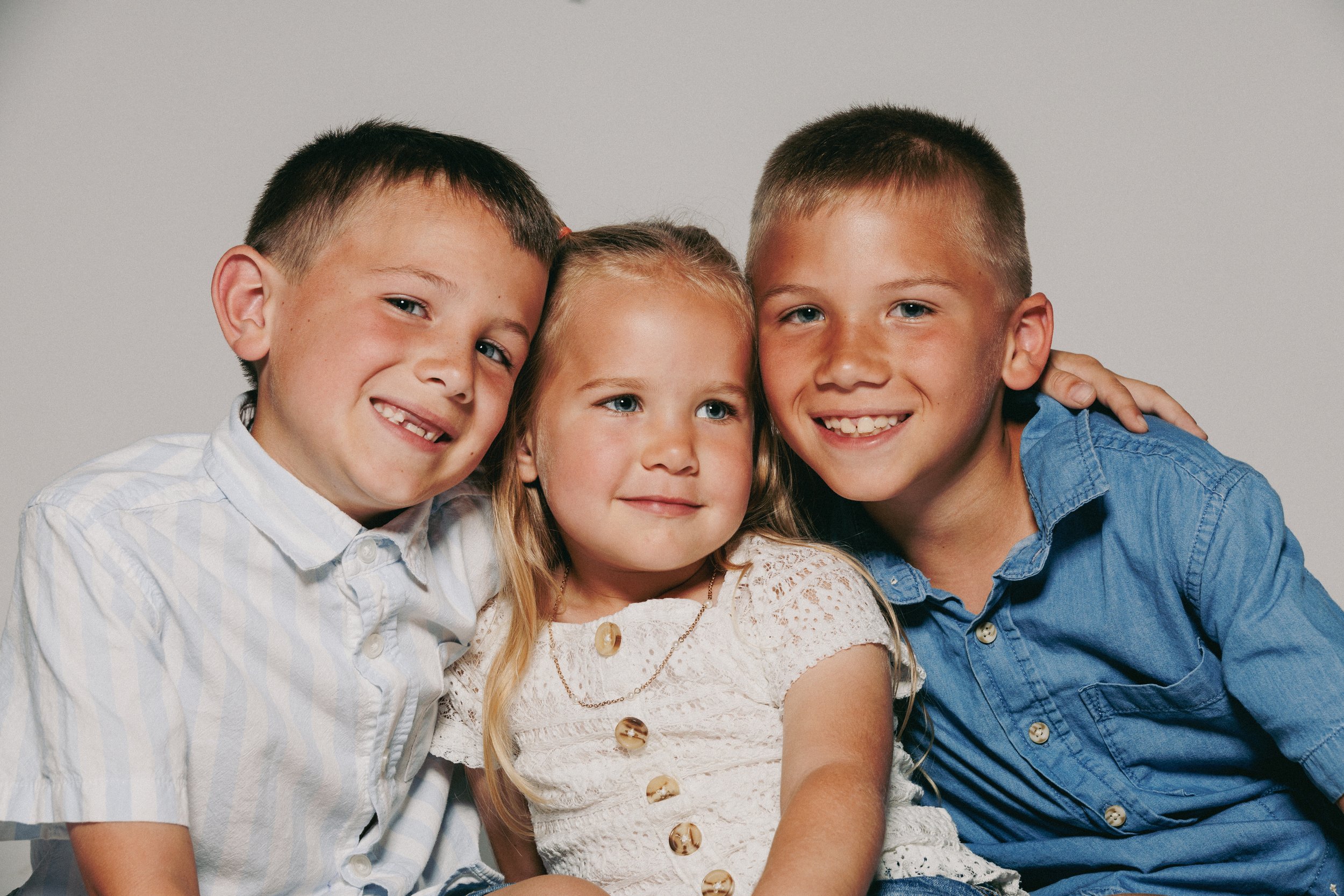 Group of four children, two boys and two girls, hugging each other and smiling at the camera, against a plain gray background.