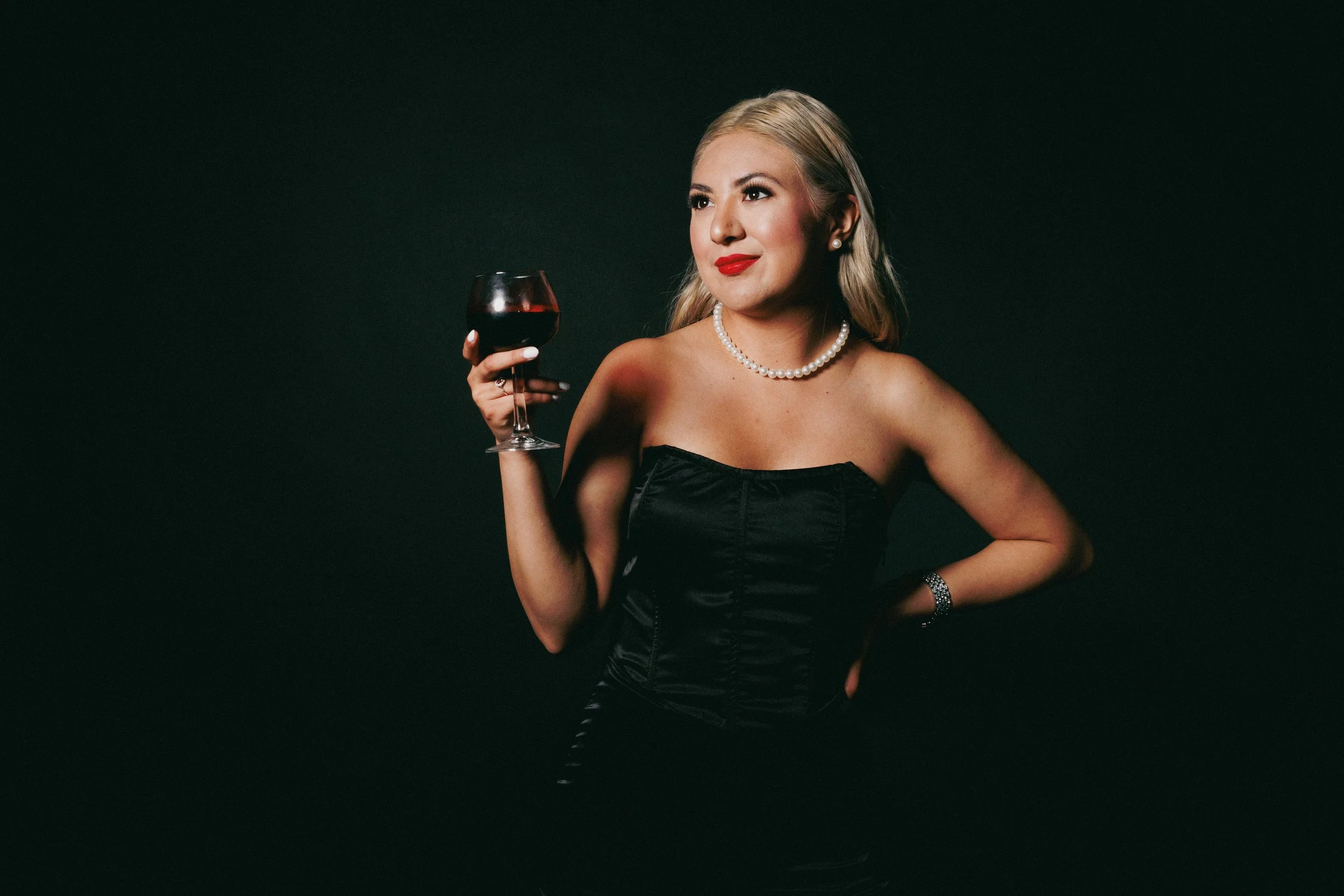 A woman with blonde hair holding a glass of red wine, wearing a black strapless dress, pearl necklace, earrings, and a bracelet, against a dark background.