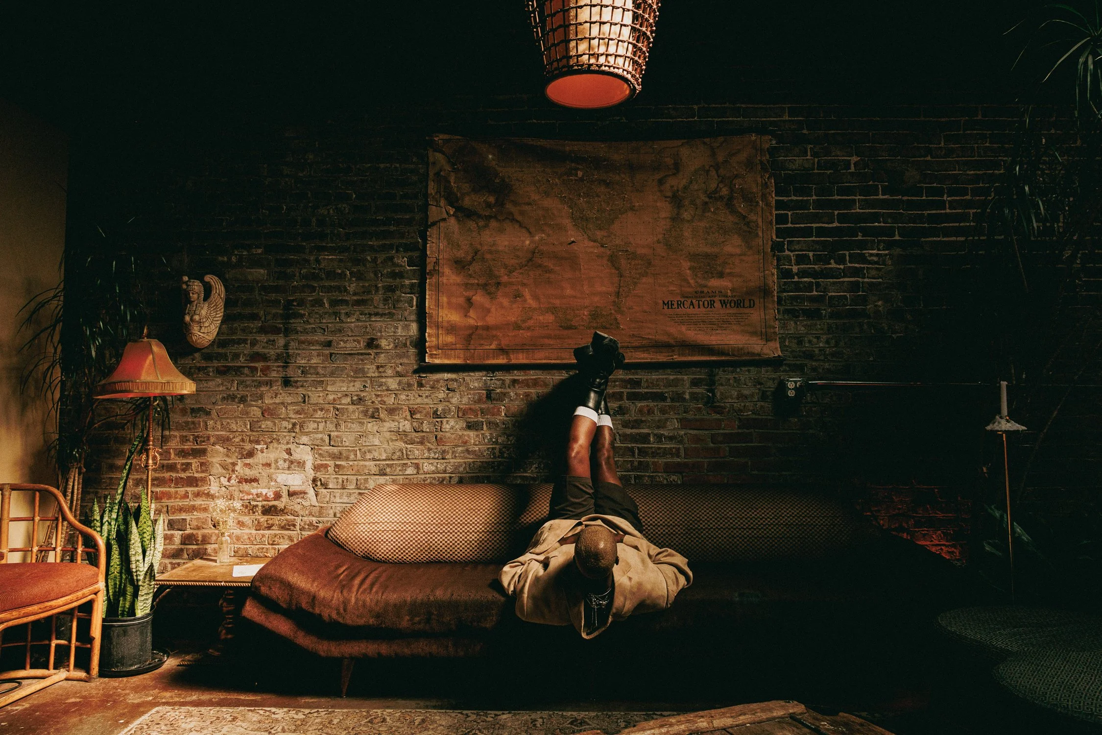 A person lying on a brown couch with their legs up against a brick wall, wearing beige pants and a jacket. There are patterned cushions, a vintage lamp, and some plants in the room. A large world map is on the brick wall.