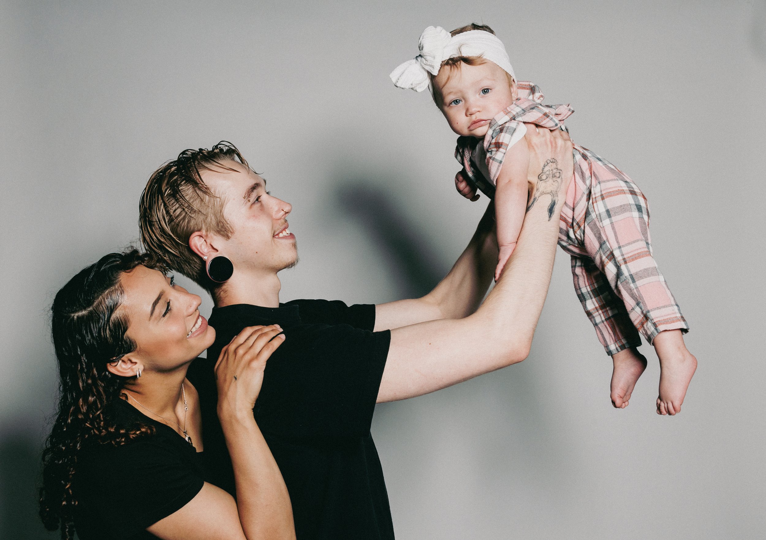 Three young adults are smiling as they hold a small child between them. The child, wearing a plaid dress and a headband, looks at the camera with a neutral expression. The background is plain and gray.