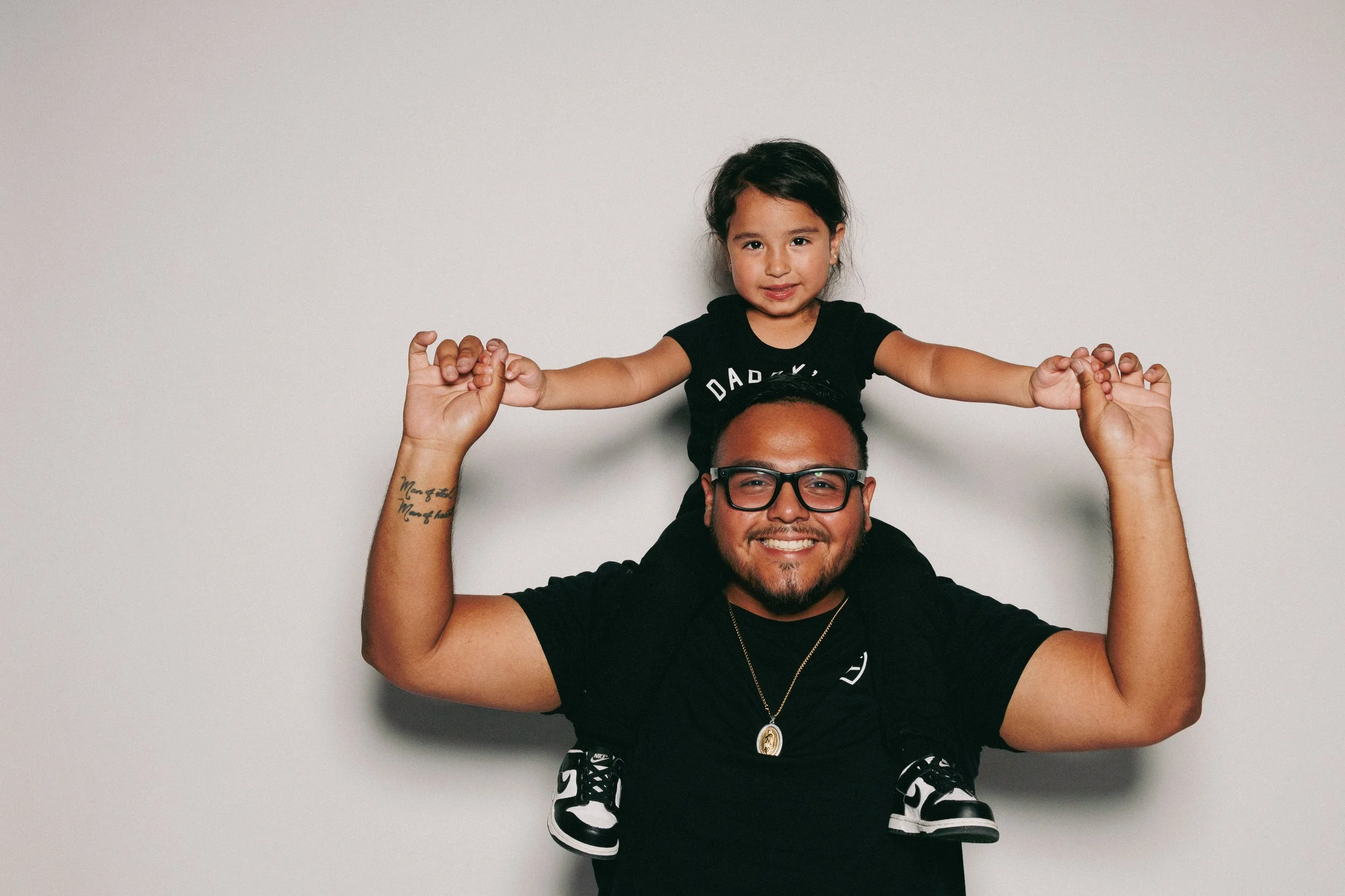 A man with glasses and tattoos on his arm is holding a young girl on his shoulders, both smiling, against a plain light gray background.