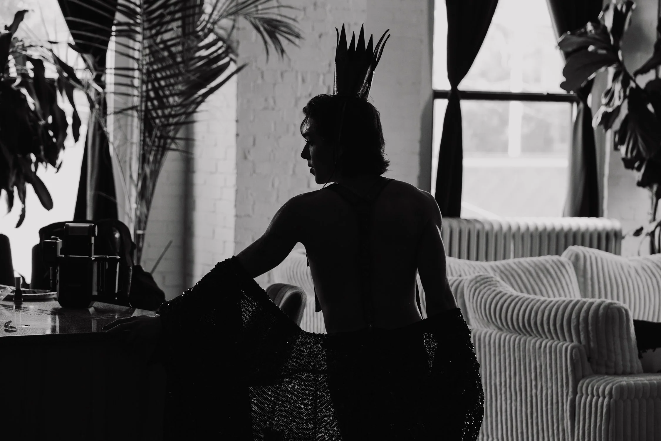 A silhouette of a woman with a headpiece standing by a table filled with makeup and accessories, indoors with large windows and potted plants.