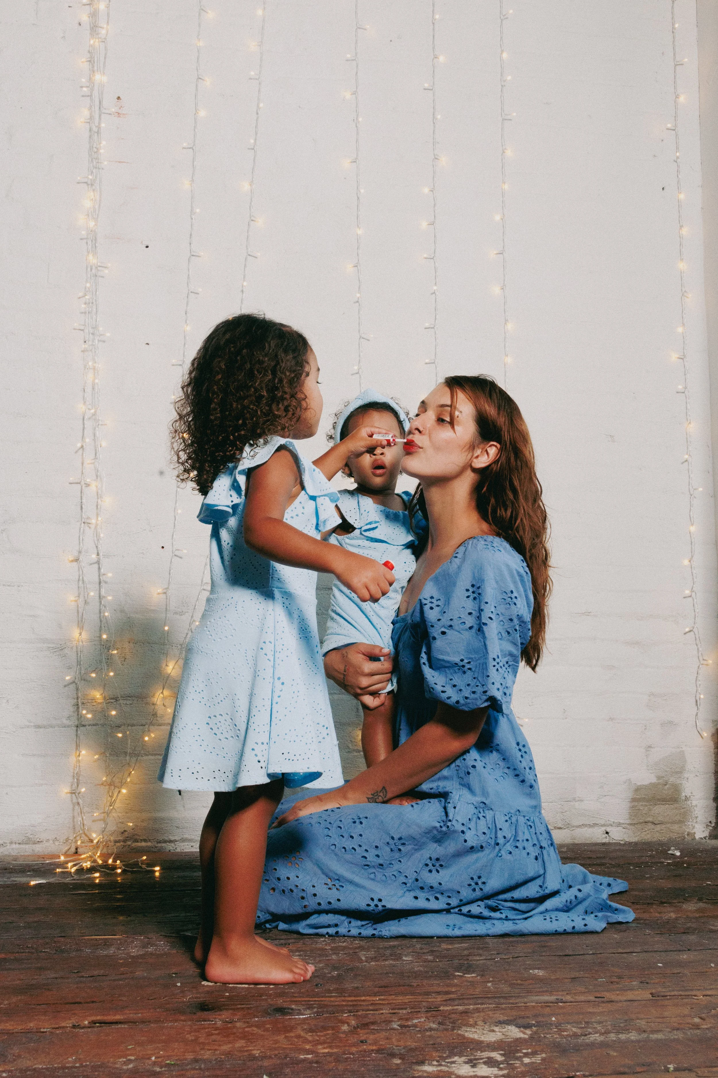 A woman with curly hair and two young girls in blue dresses, one of whom is putting lipstick on the woman's lips, standing in front of a white wall decorated with string lights. The woman is kneeling on a wooden floor.