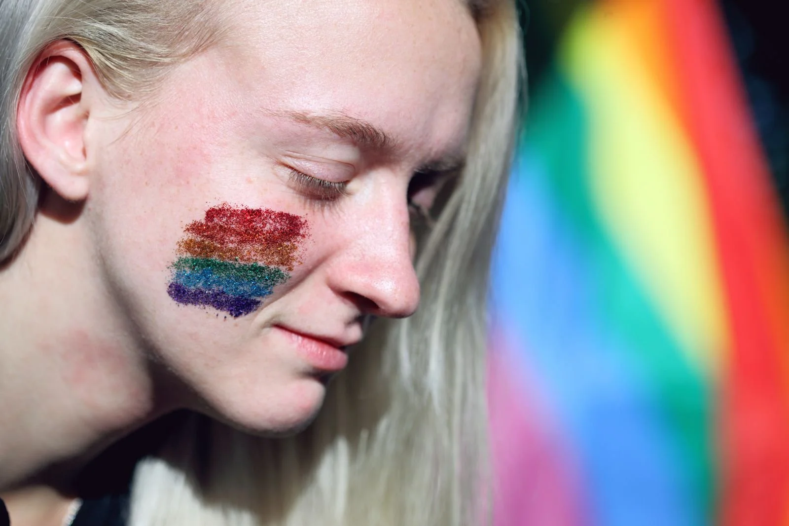 Close-up of a person with blonde hair, closed eyes, and a glittery rainbow flag painted on their cheek, symbolizing LGBTQ+ healing and trauma support