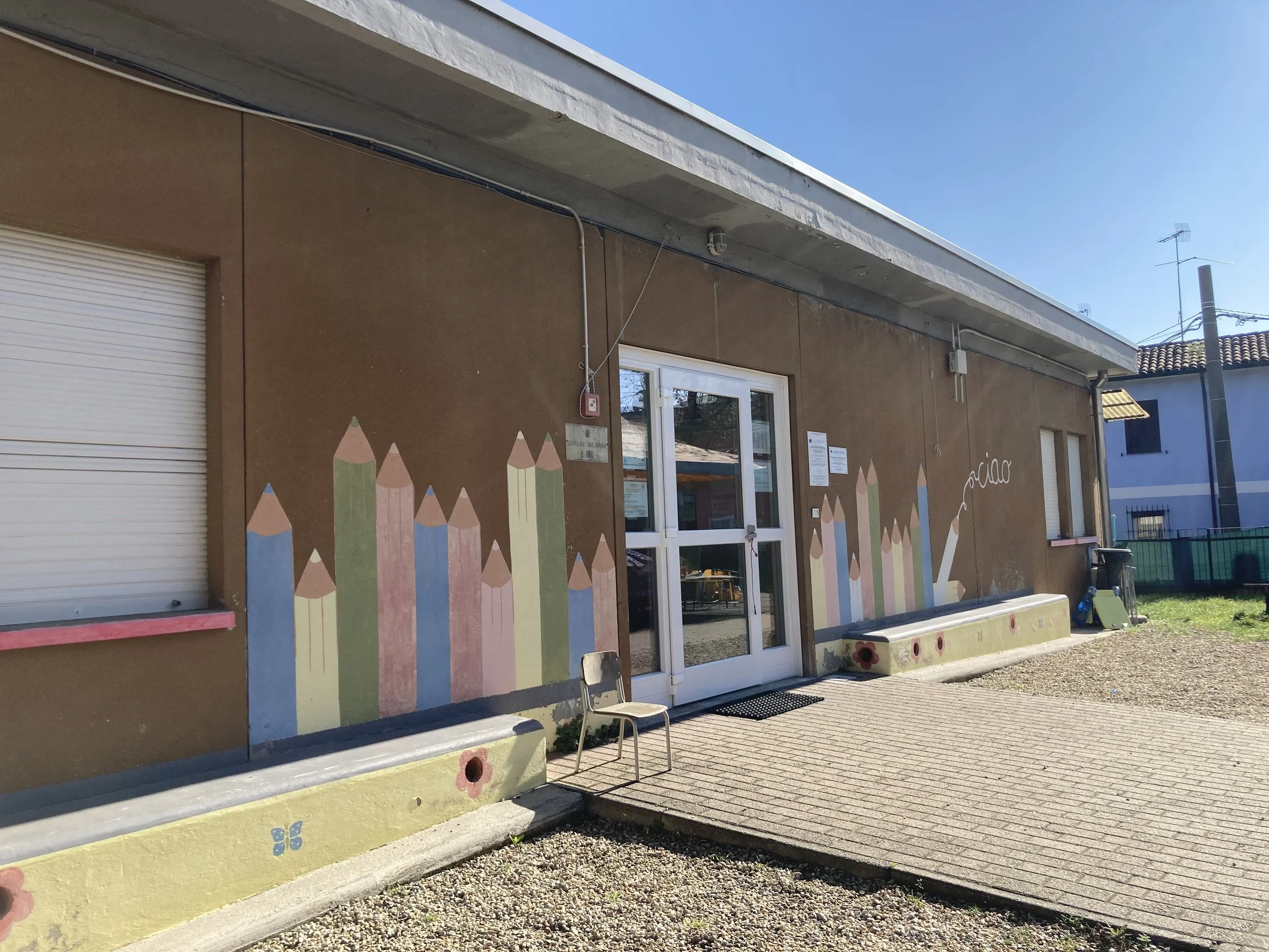 Facciata di un edificio con decorazioni di matite colorate disegnate sulla parete esterna. Ci sono due sedie di plastica davanti alla porta d'ingresso, con un ambiente scolastico o per bambini.