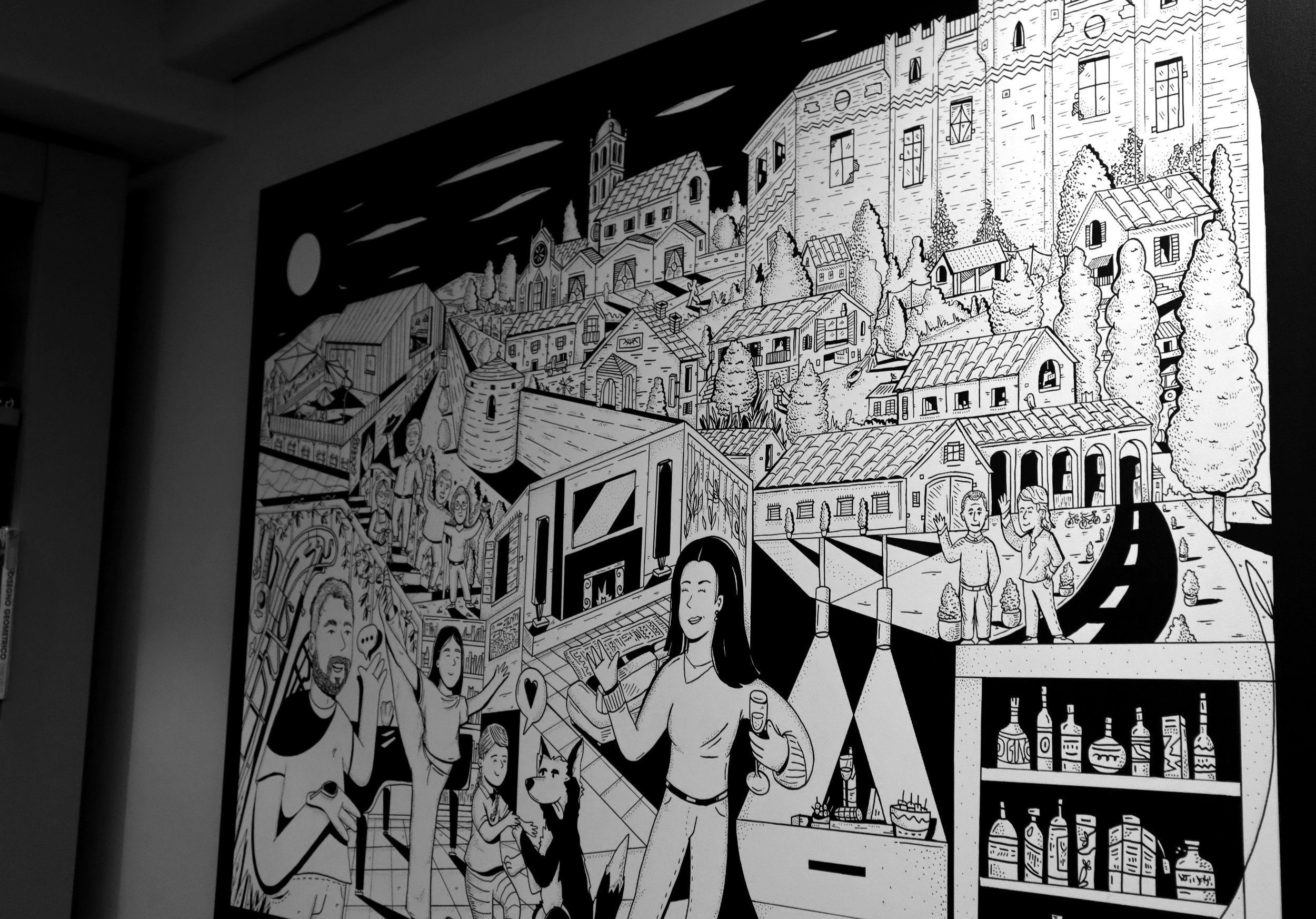 Mural nero e bianco che rappresenta un paesaggio urbano con case, alberi, una chiesa e persone che socializzano, con elementi decorativi come bottiglie, cibo e un cane.