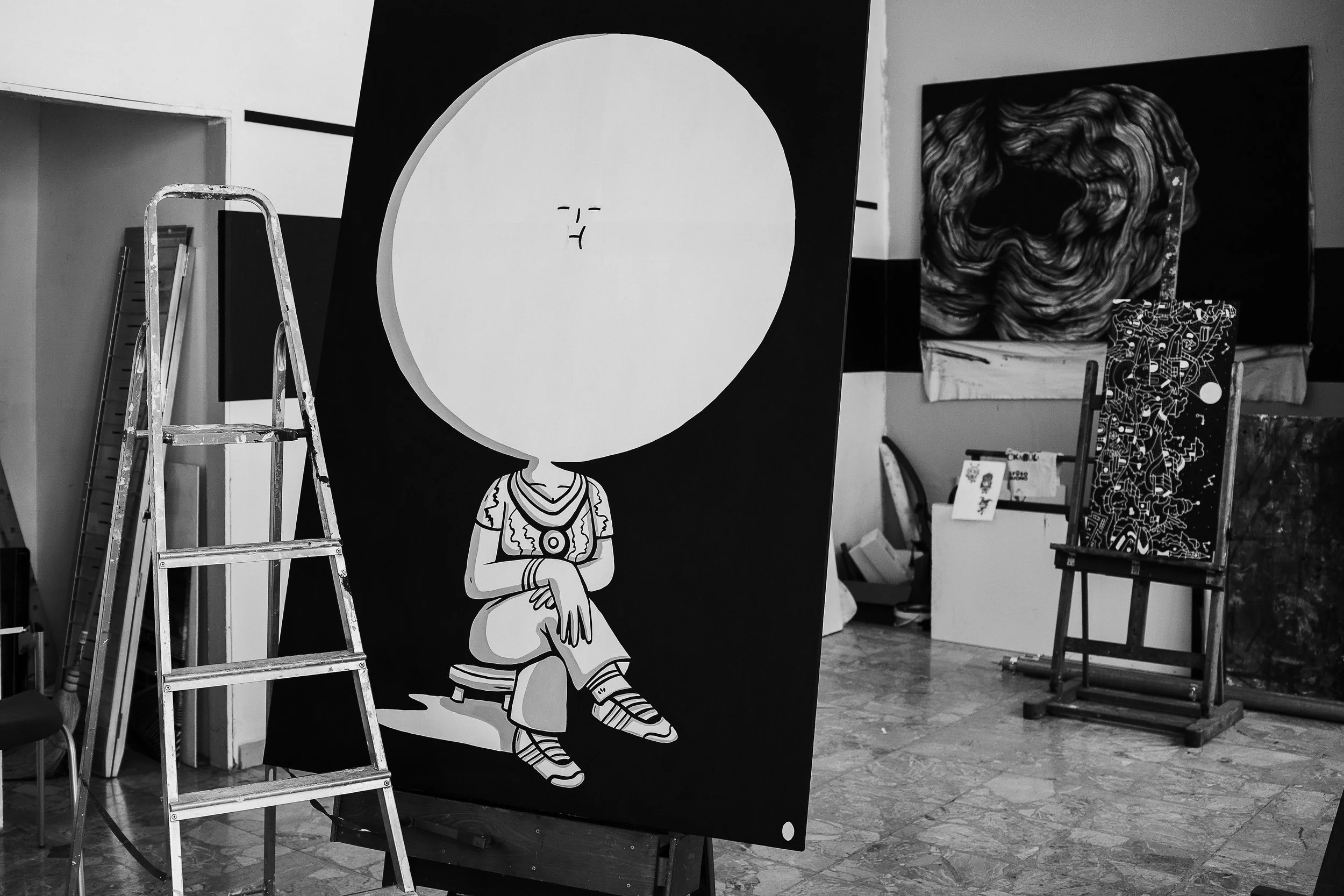 Un'opera d'arte in bianco e nero raffigura una figura umana con un grande cerchio sopra la testa, seduta in posa con le gambe incrociate, abbigliamento e acconciatura stilizzati. Lo sfondo mostra altri quadri e strumenti artistici.