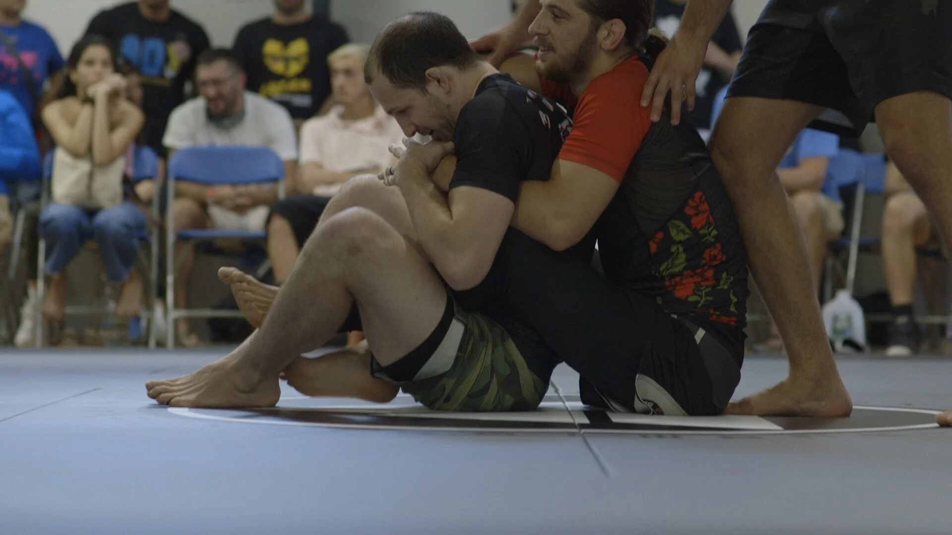 Two men grappling during a Brazilian Jiu-Jitsu match with spectators watching in the background.