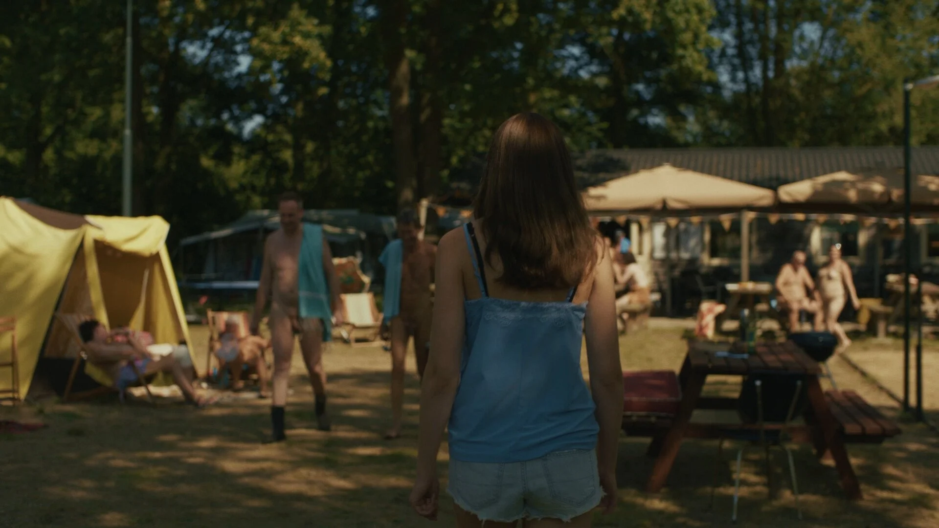 A girl with shoulder-length brown hair wearing a blue tank top and denim shorts stands outdoors, facing a group of people enjoying a poolside gathering under umbrellas and tents, with lush green trees in the background.