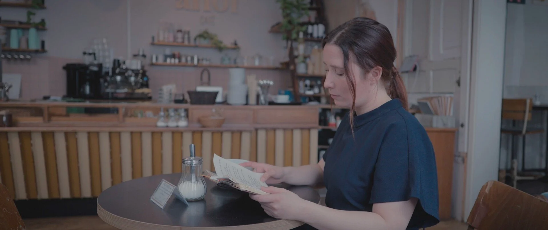 A woman in a dark blue shirt sitting at a round table in a cafe, reading a menu. There are sugar and milk containers on the table, with a cozy cafe interior in the background.