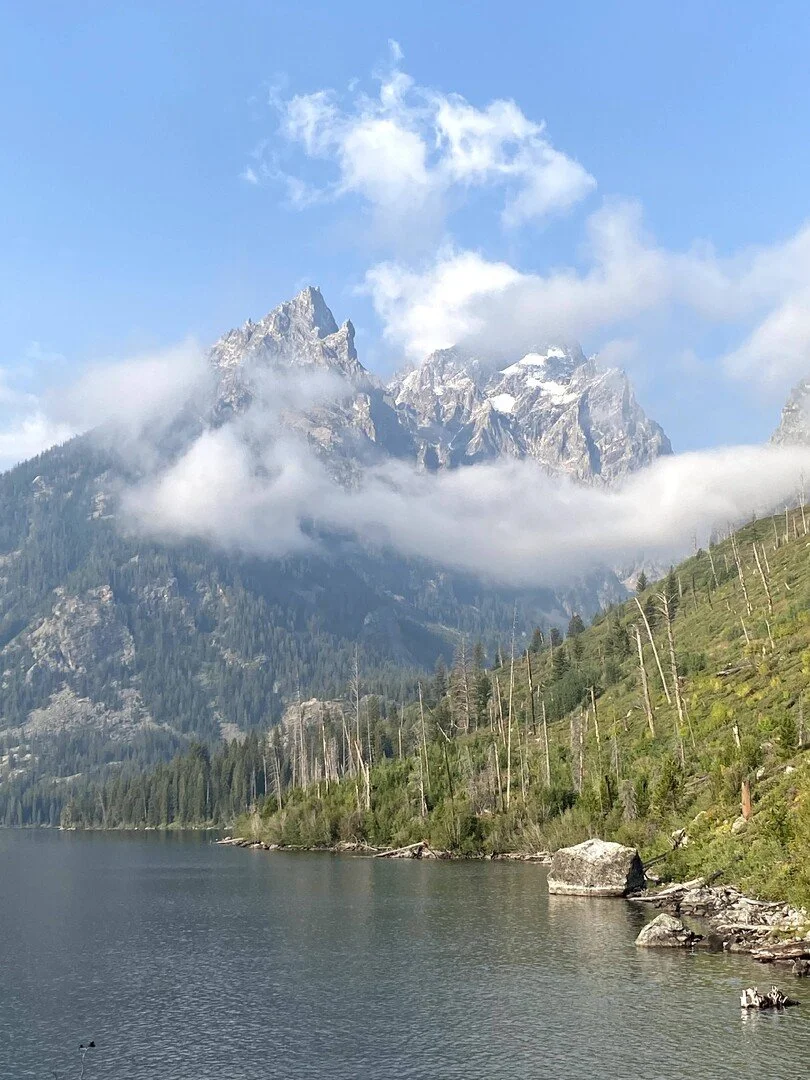 Mountain lake with evergreen trees and rocky shoreline, snow-capped peaks in the background, and a partly cloudy blue sky.