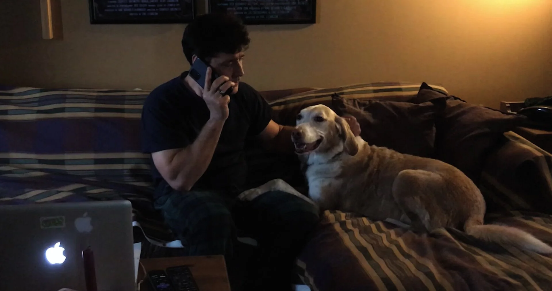 A man sitting on a bed talking on a cell phone with a golden retriever lying beside him. There is a laptop, TV remote, and other items on a table nearby.