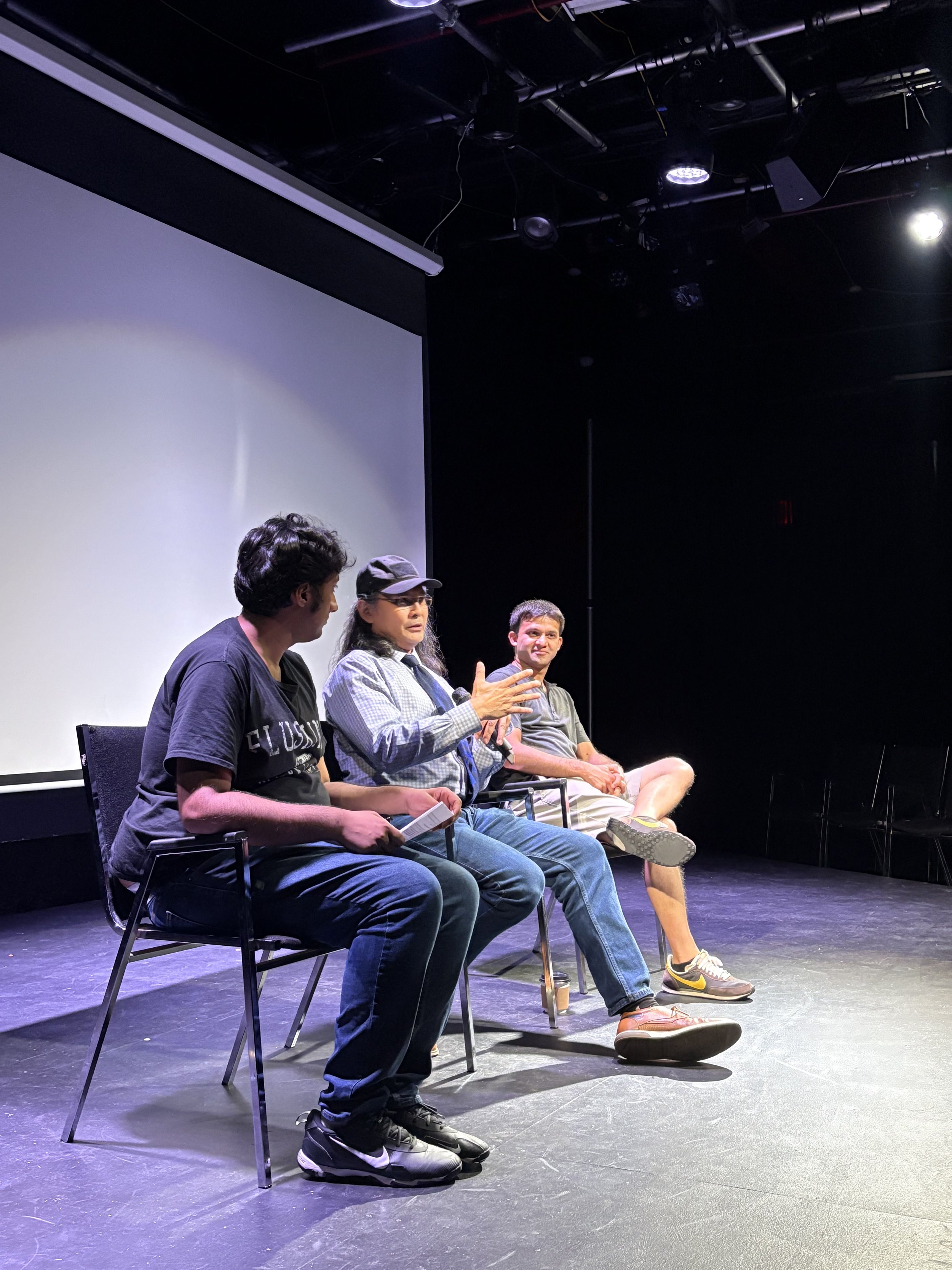 Three people sitting on chairs in a dark theater or conference room, engaged in a discussion, with a plain white screen behind them.