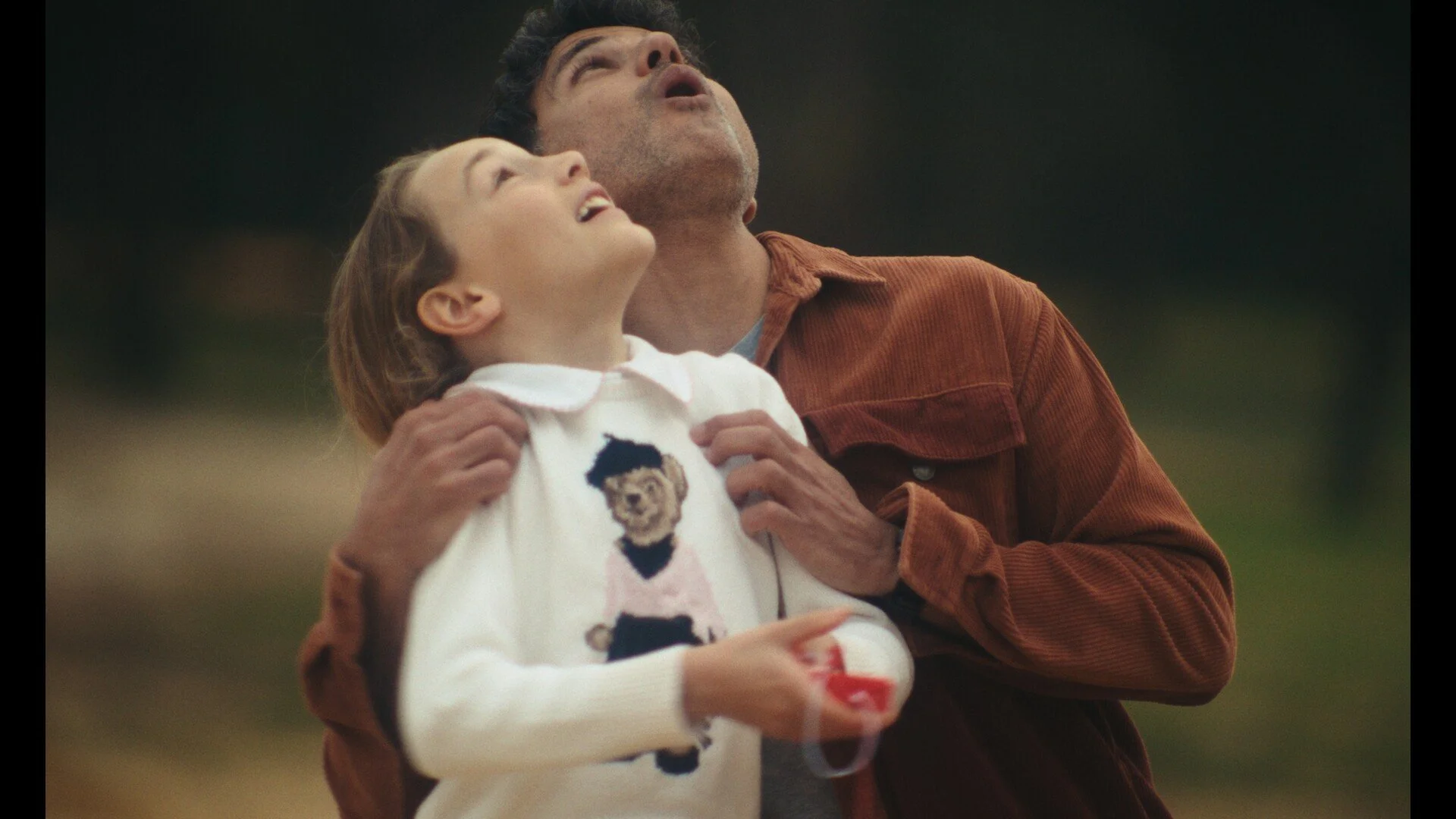 A man and a young girl outdoors looking up at the sky with puzzled or surprised expressions, the girl holding a transparent object.