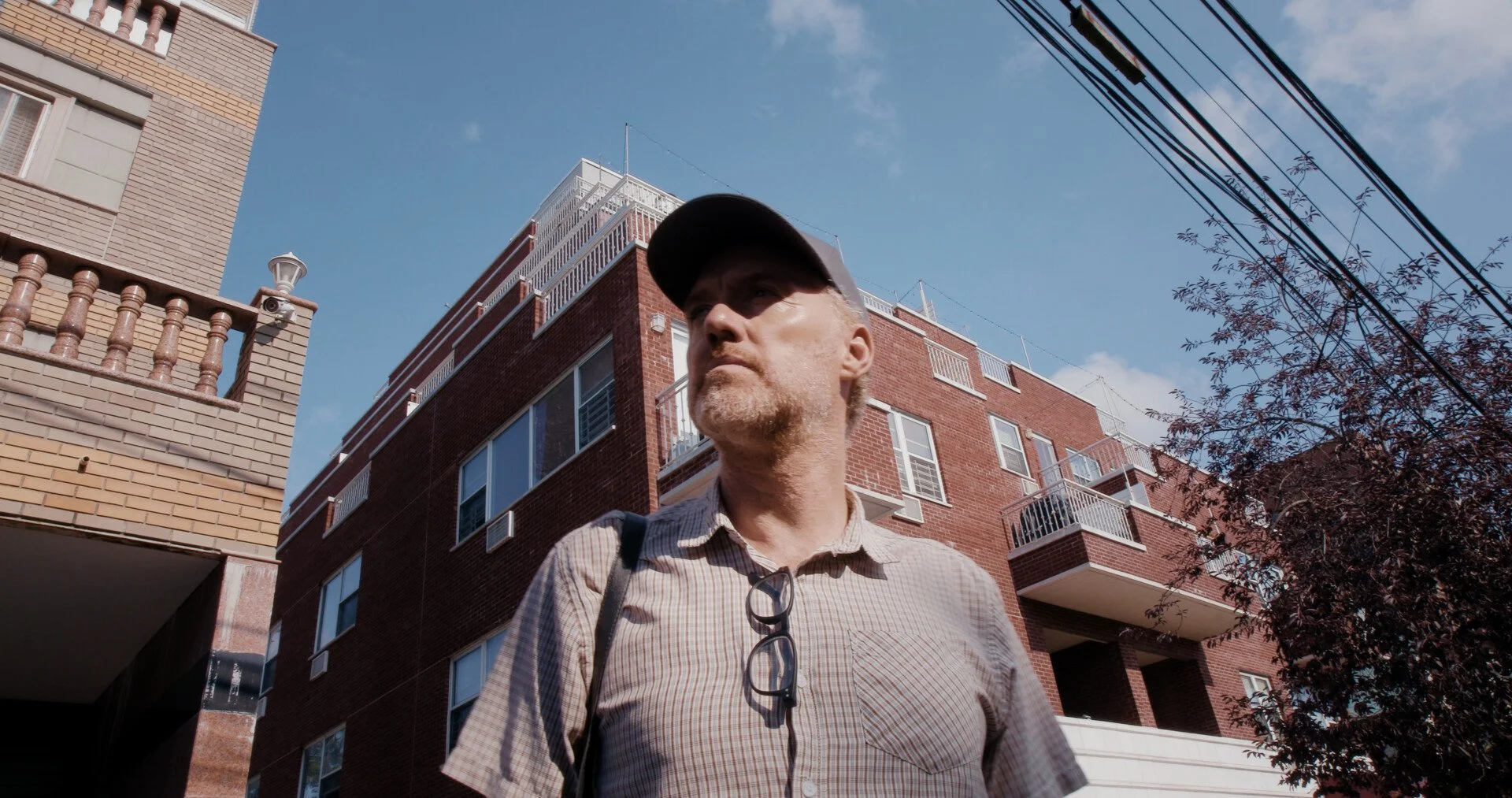A man wearing a cap, with glasses hanging from his shirt, stands outside in front of a multi-story brick building under a blue sky with power lines overhead.