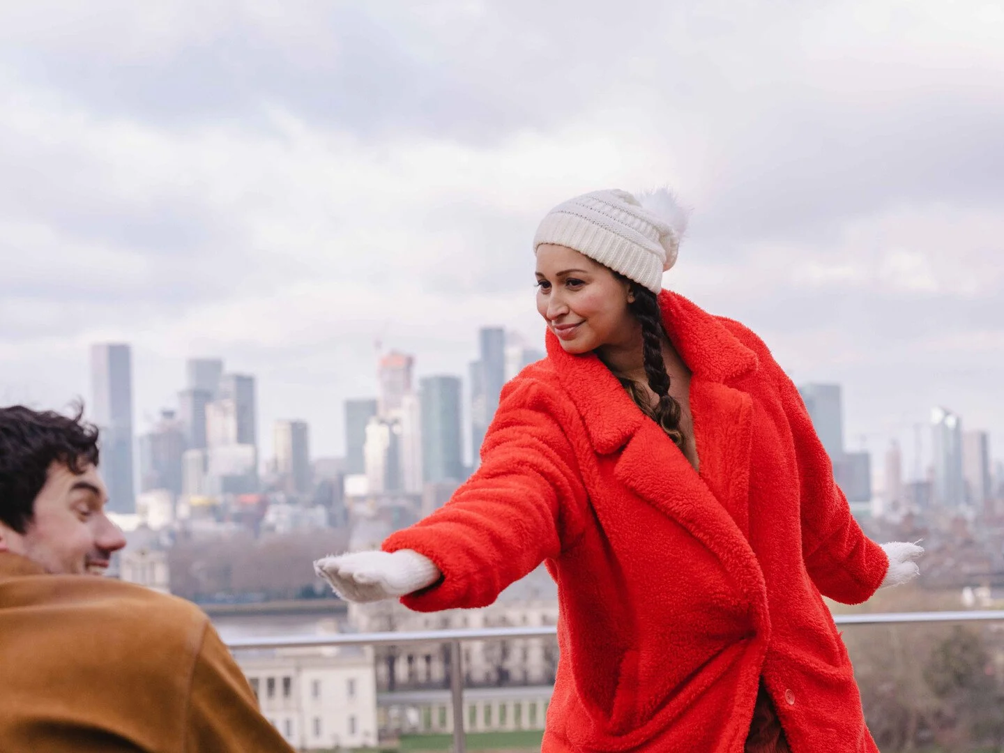 A woman in a red coat and white winter hat offering her hand to a man in a brown jacket on a rooftop with a city skyline in the background.