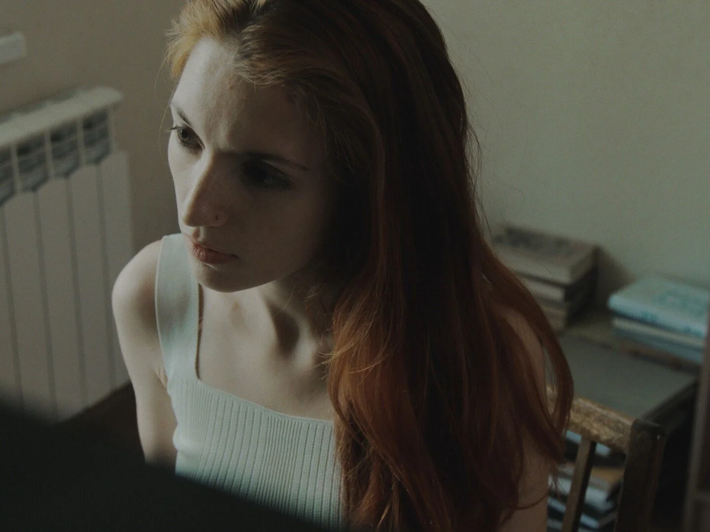 A young woman with long red hair and a nose piercing, wearing a white tank top, looking to her left in a dimly lit room with a stack of books and a chair in the background.