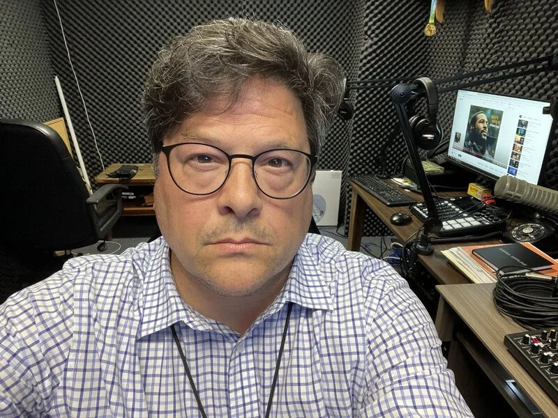 A man with glasses and brown hair taking a selfie in a recording studio, surrounded by soundproof foam panels, audio equipment, a computer monitor displaying a person speaking, and a microphone.