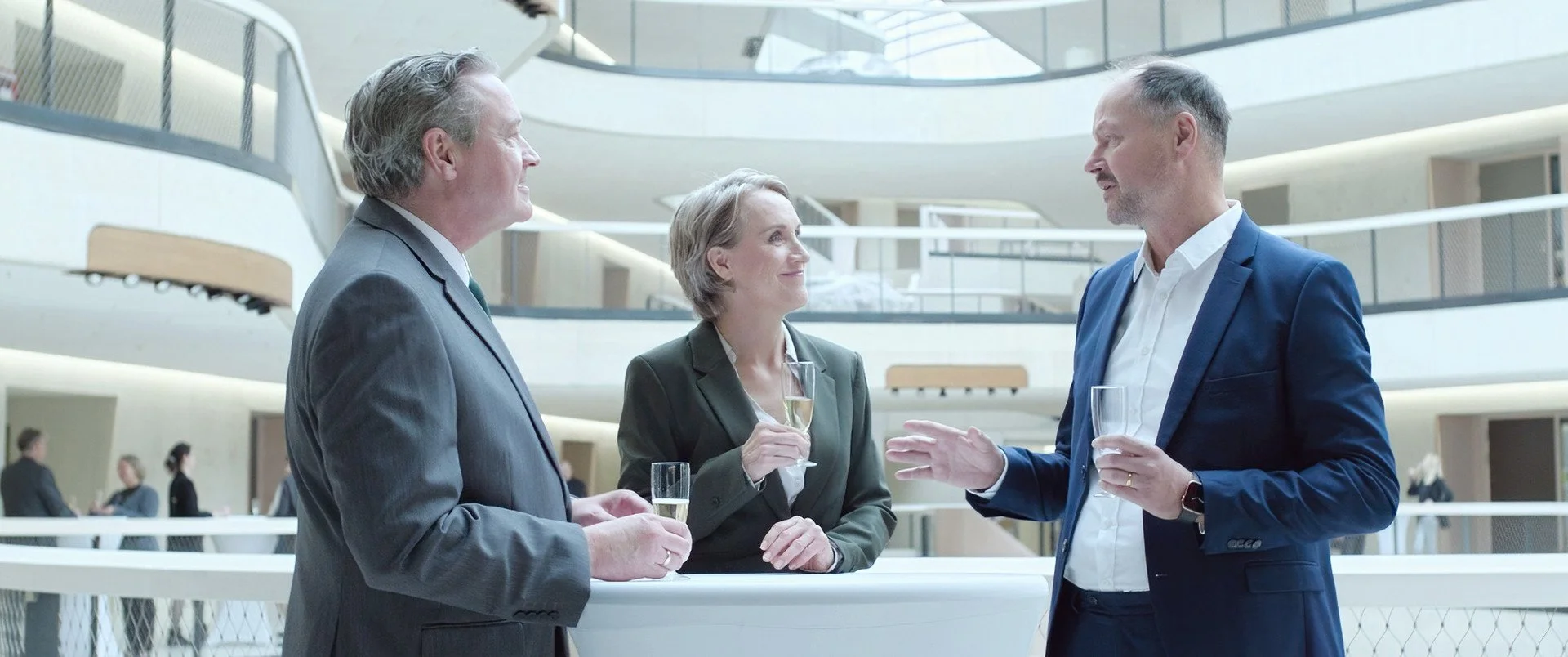 Three business professionals in suits having a conversation with champagne glasses at a modern office building reception area.