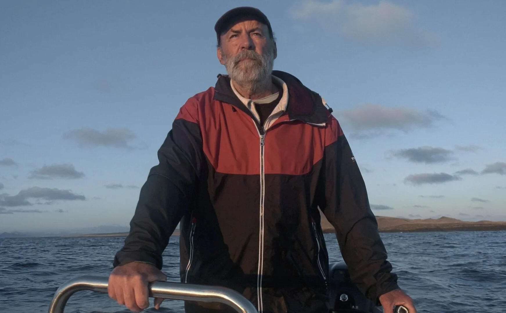 An older man with gray hair and beard on a boat, holding the rail, wearing a red and black jacket and a dark baseball cap, with water and distant landscape behind him during sunset or sunrise.