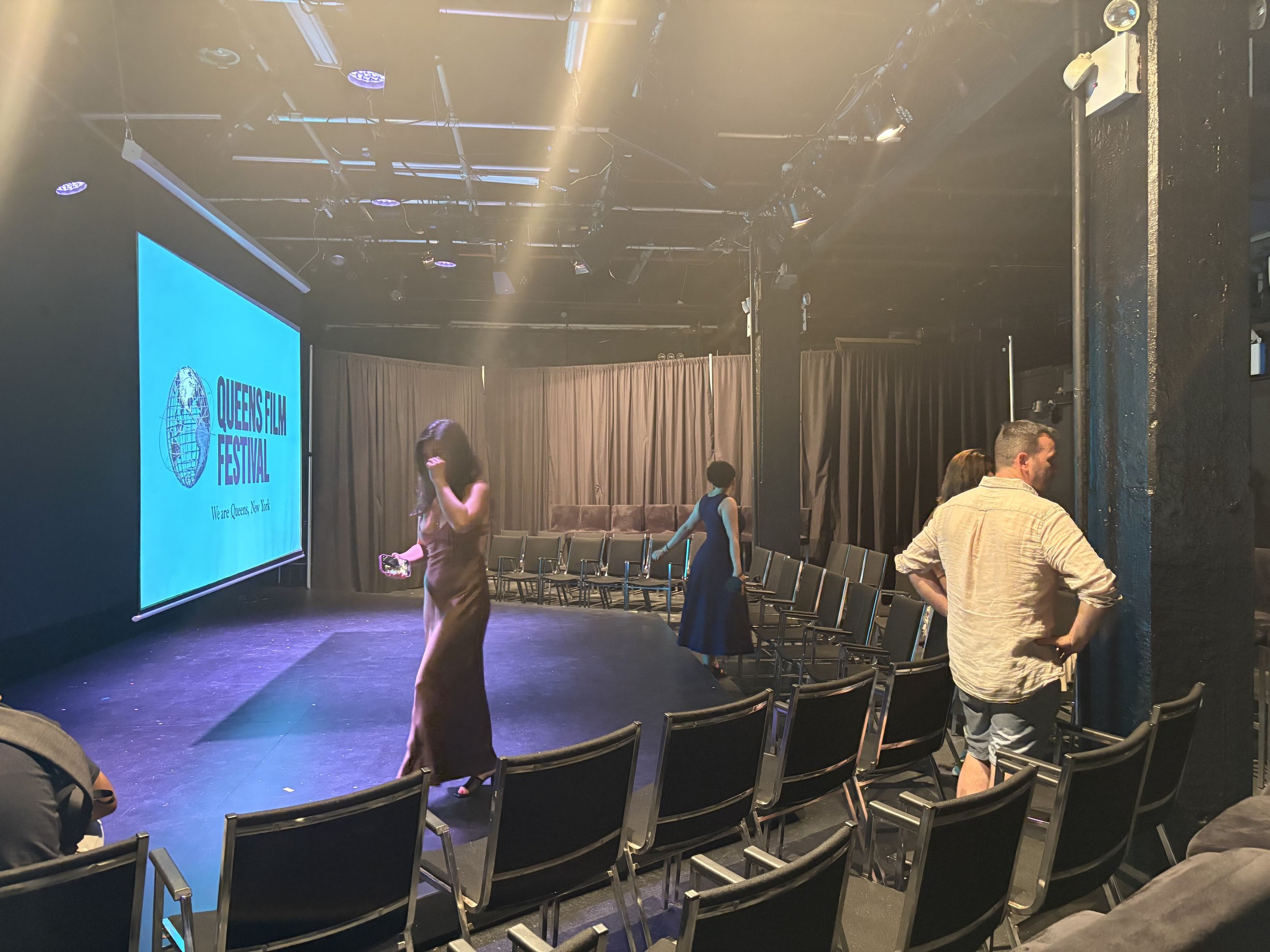 An empty stage with chairs, a large screen displaying 'Queens Film Festival,' and a few people standing and walking around the seating area.
