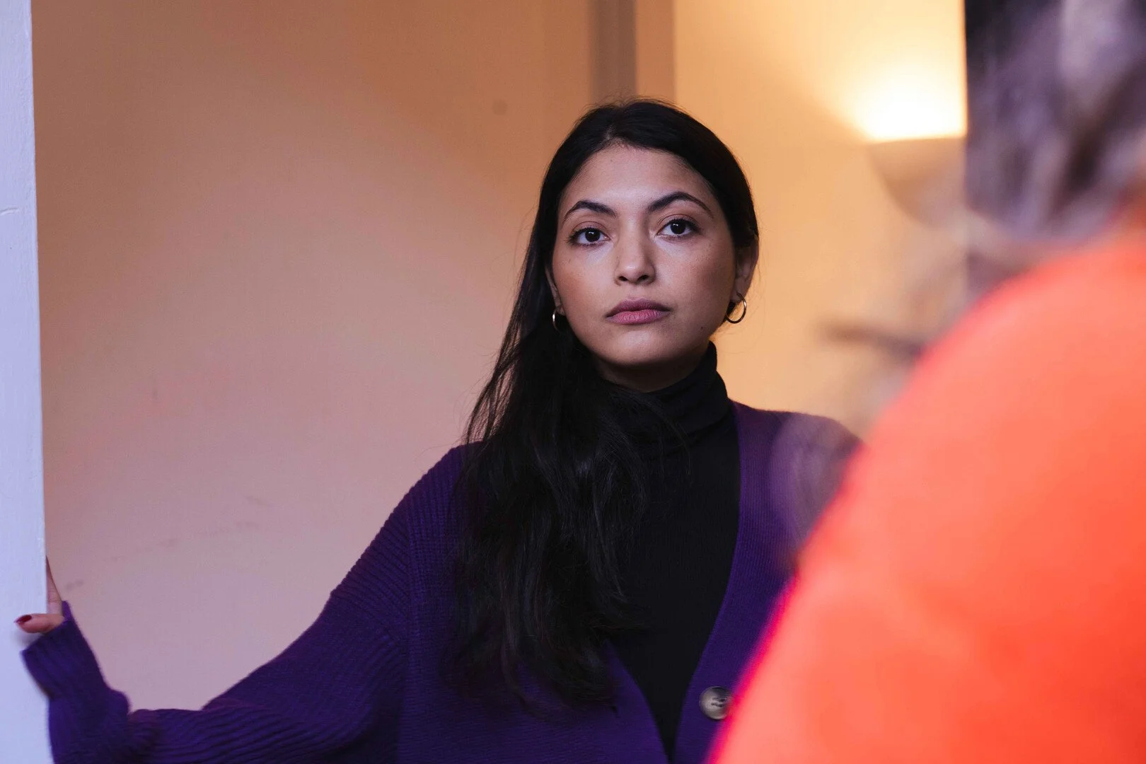 A young woman with long dark hair and hoop earrings standing in a doorway, looking directly at the camera, wearing a black turtleneck and purple cardigan, with warm indoor lighting.