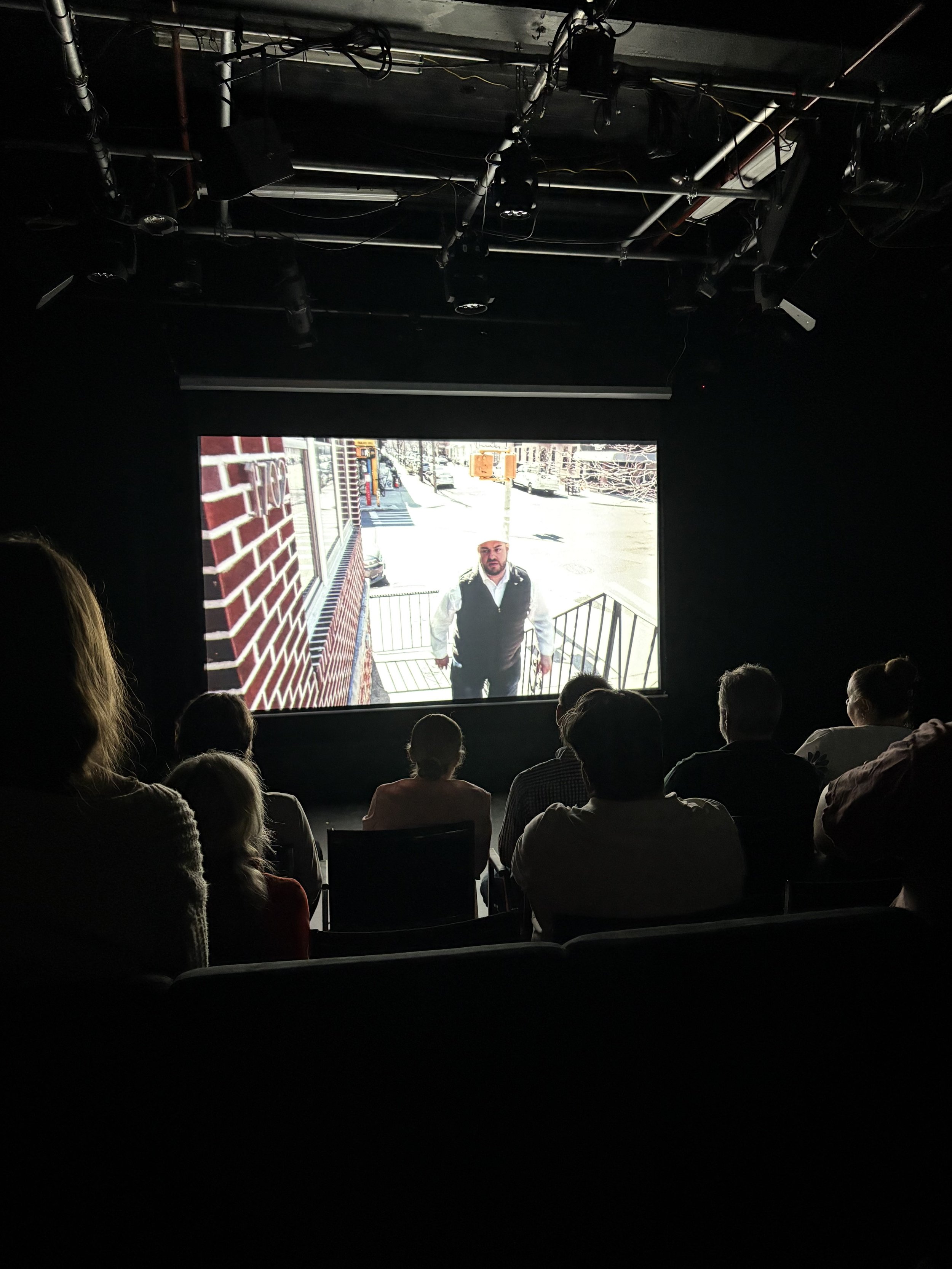 Audience watching a movie or presentation in a dark theater with a large screen showing a man standing outside on a street, near a staircase leading down from a building, with brick walls and city street background.