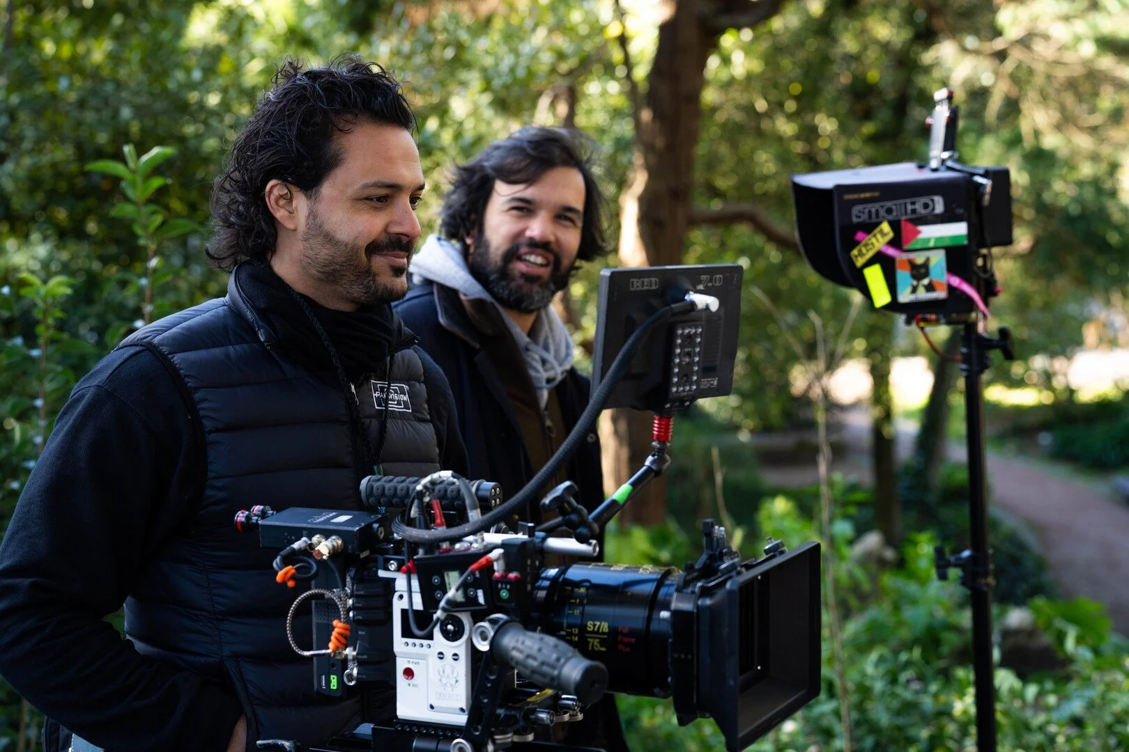 Two men operating a professional film camera in an outdoor setting with green trees and plants.