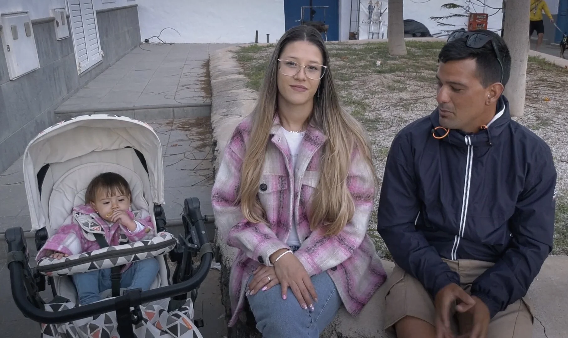 A woman with long hair and glasses sitting on a bench with a man, both looking slightly to the side. There is a young girl sitting in a stroller on the woman's left.