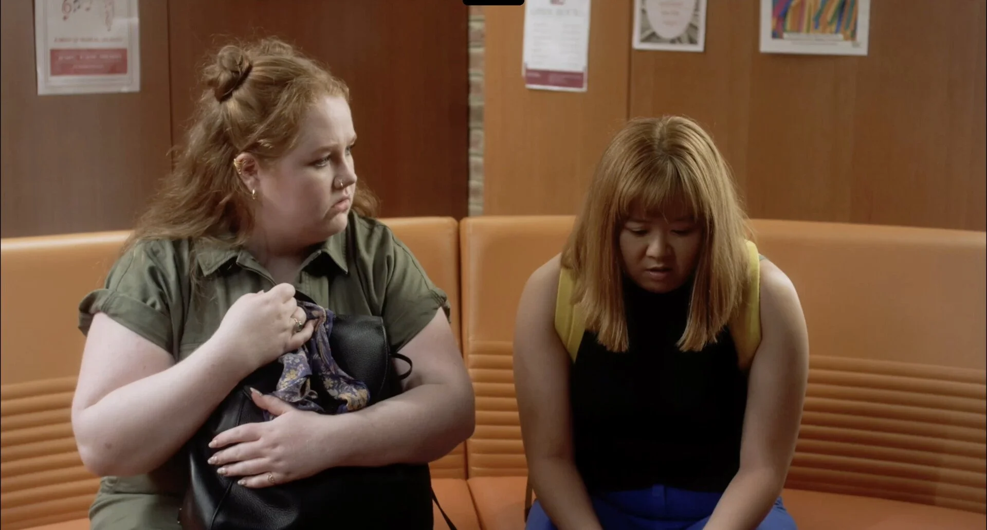 Two young women with red hair sitting on a brown cushioned bench in a waiting area, one holding a purse and looking at the other, who appears upset or deep in thought.