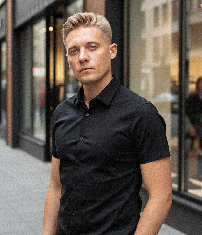 A young man with blonde hair styled back, light skin, wearing a black button-up shirt, standing outside in an urban setting with glass storefronts and blurred city street in the background.