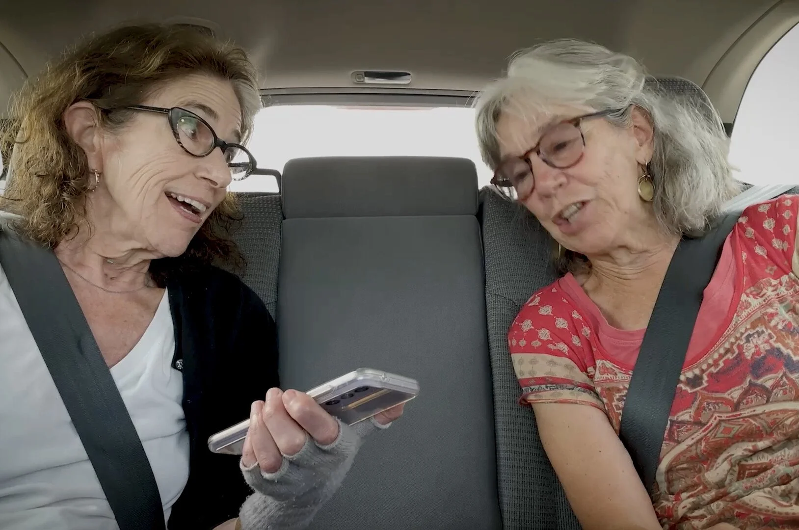 Two older women with glasses sitting in the back of a car, looking at a smartphone.