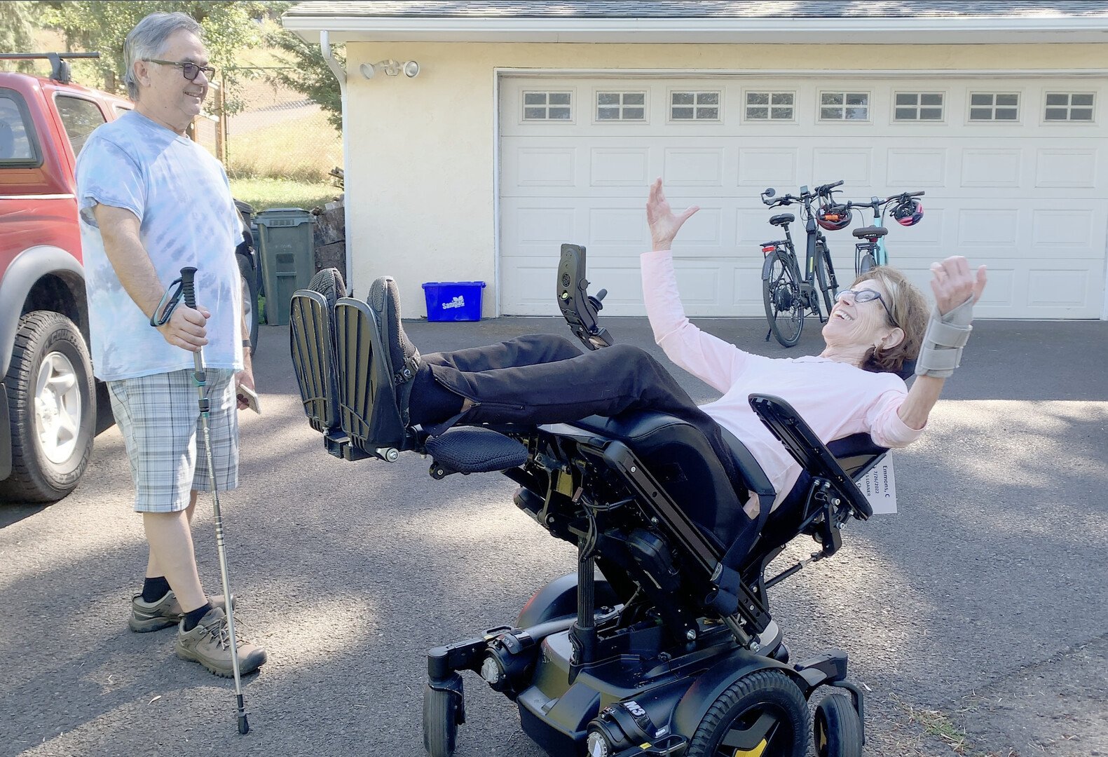 A woman in a powered wheelchair and a man standing beside her in a driveway, smiling and appearing to have fun. The woman is laughing and raising her arms, with bikes and a garage in the background.