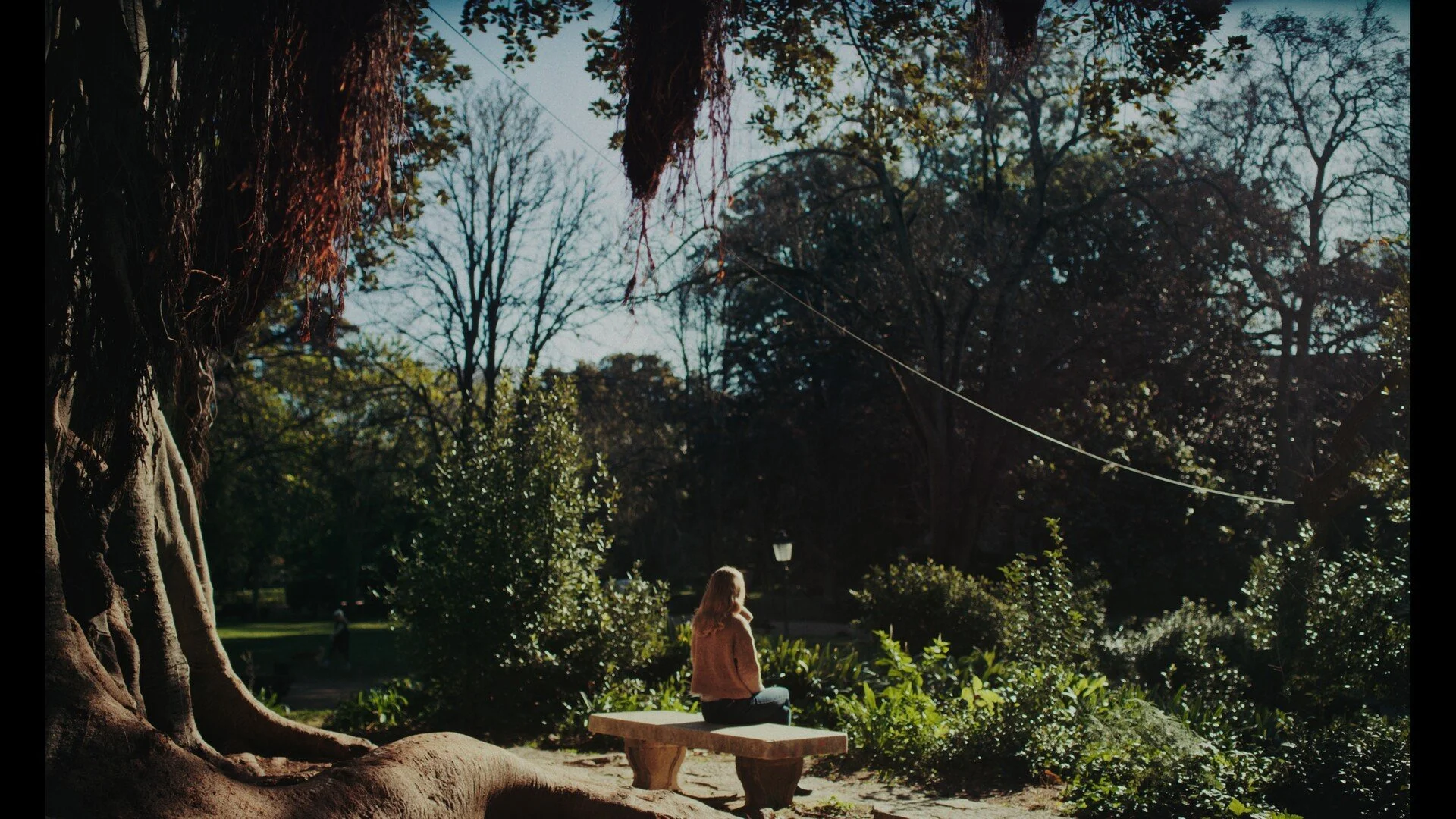 A woman with long blonde hair, wearing a brown coat, sits on a stone bench in a park surrounded by trees and lush green bushes during daytime. The sunlight filters through the trees, creating a peaceful atmosphere.