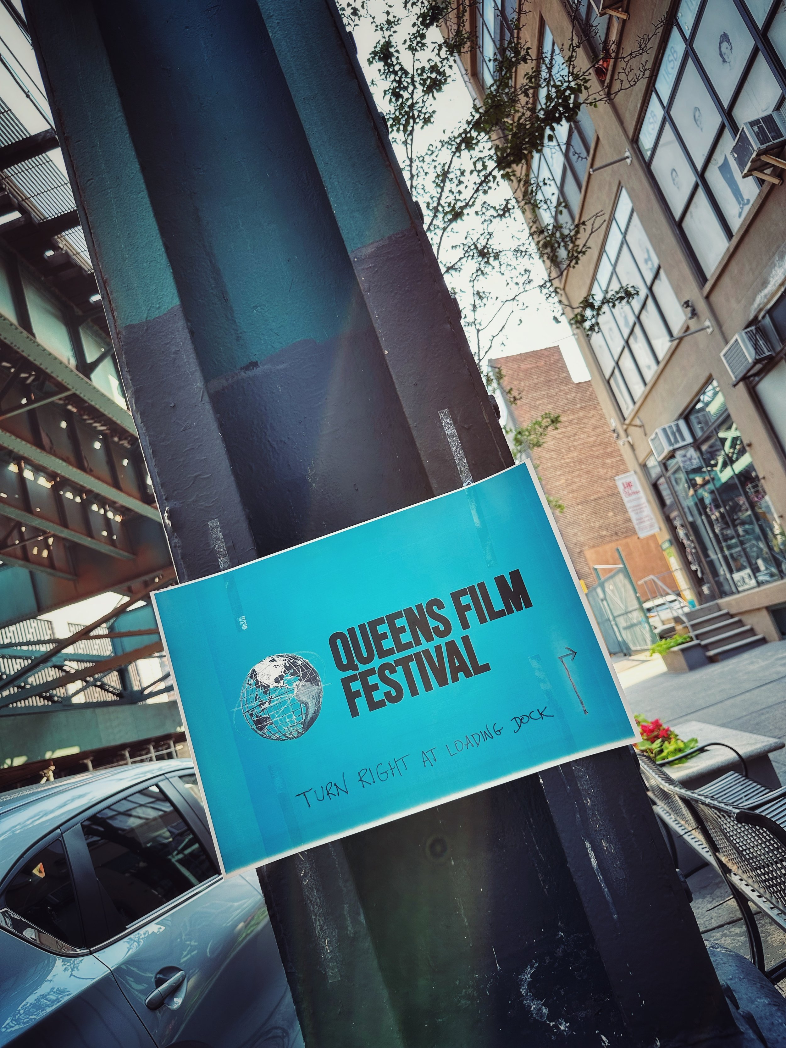 Sign for Queens Film Festival with globe icon, attached to a black pole on a city sidewalk, buildings and trees in the background