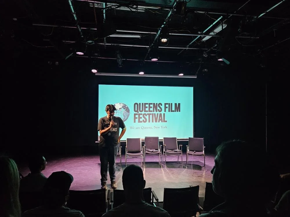 A person speaking on stage at the Queens Film Festival, with a large screen behind displaying the festival's logo and text.