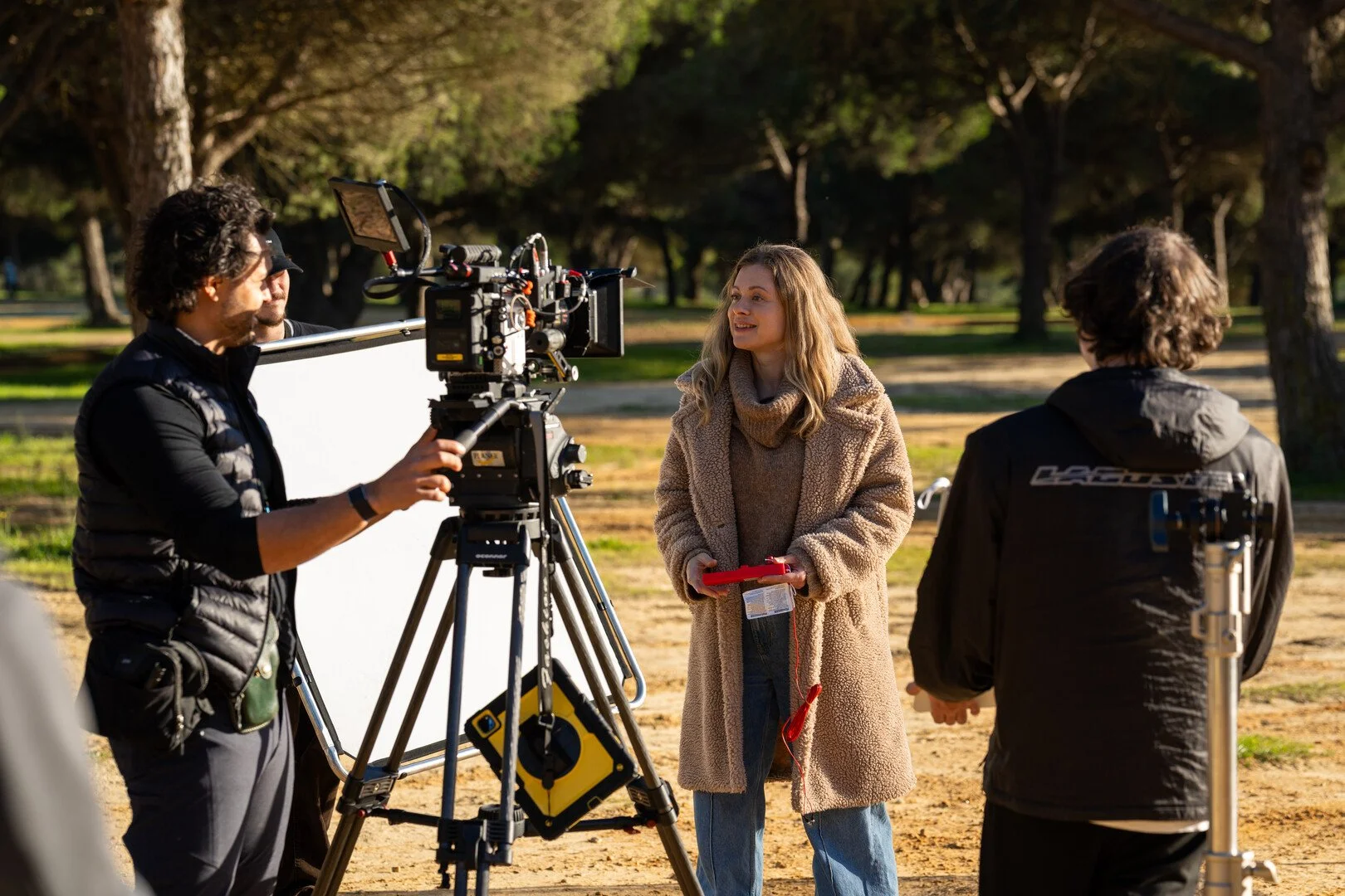 Filmmakers filming an interview outdoors in a park with trees in the background, a woman holding a red tablet speaking, and crew members operating camera equipment.