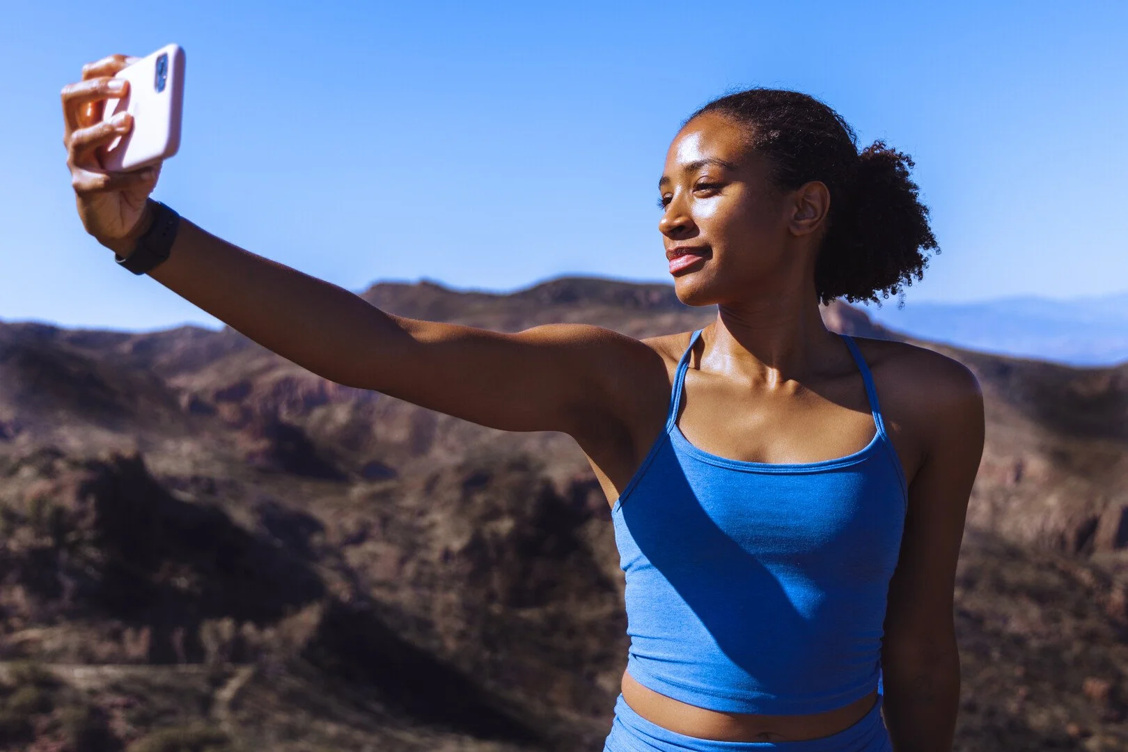Woman in blue athletic tank top taking a selfie outdoors against a mountainous landscape under a clear blue sky.