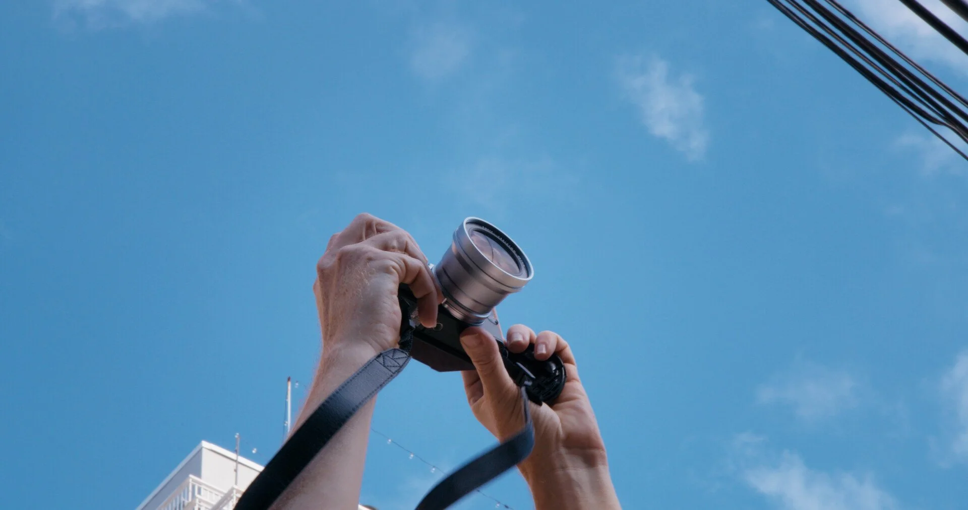 Person holding a camera pointed upward at a clear blue sky with a few clouds and power lines in the background.