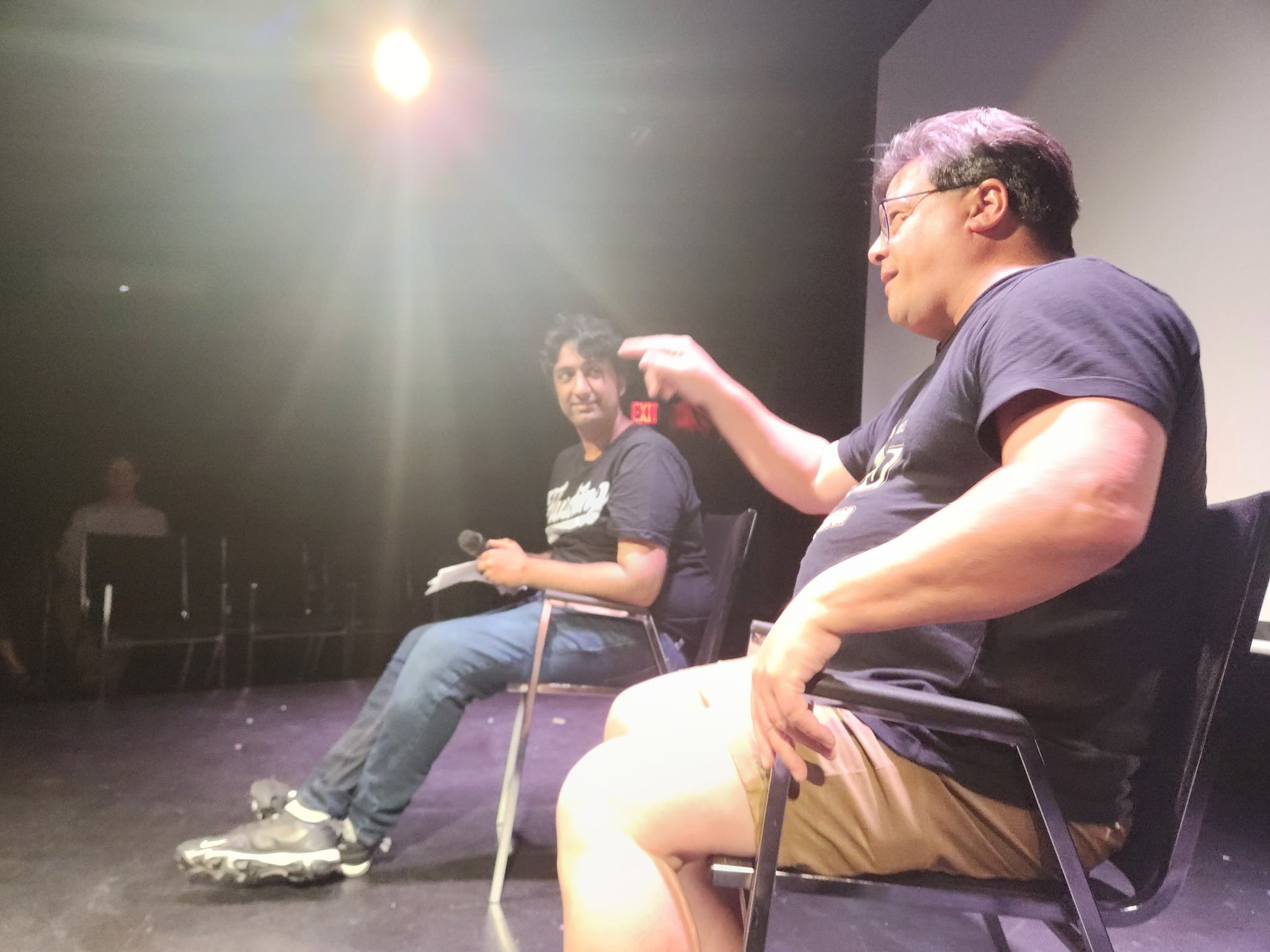 Two men are sitting on chairs on a stage. The man on the left has curly black hair, is holding a microphone and some papers, and is looking at the camera. The man on the right has glasses, short dark hair, and is gesturing with his hand. There is a bright light source overhead, and a person in the background near a wall.