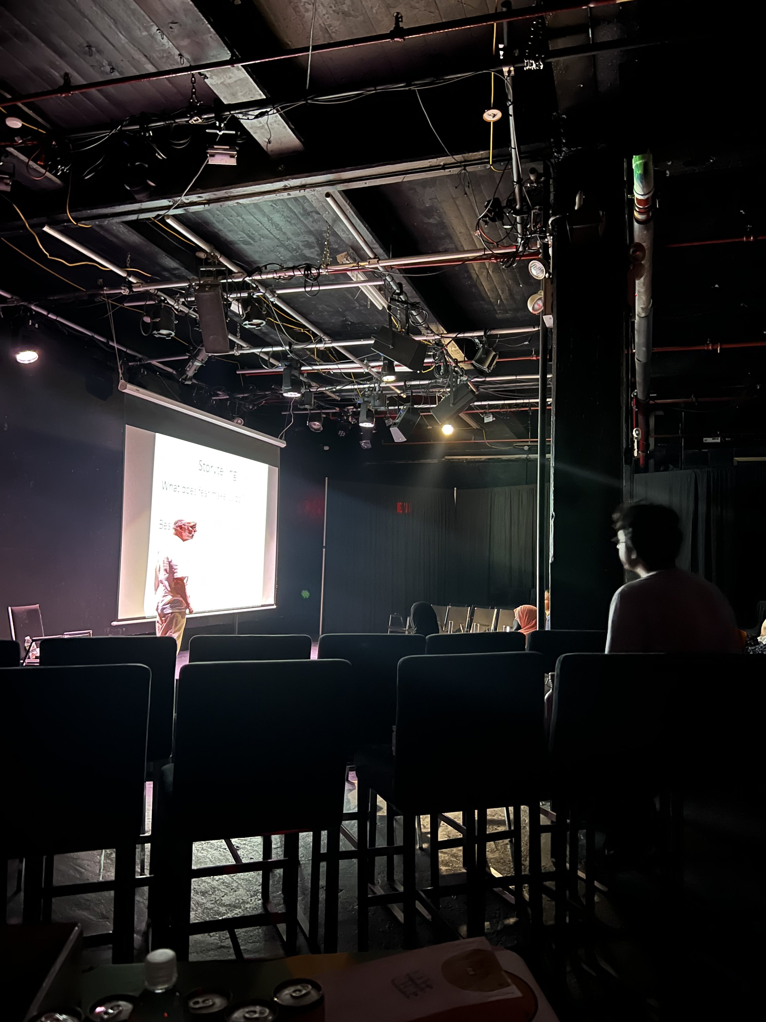 An indoor theater or presentation space with a large screen, some people seated facing the stage, and a person presenting on stage.