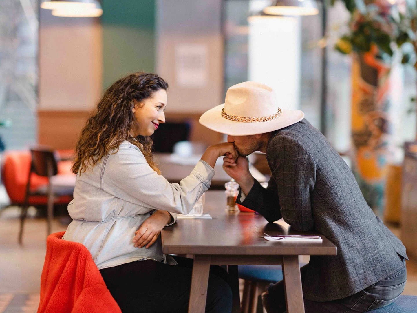 A man with a hat kisses a woman's hand at a restaurant table.