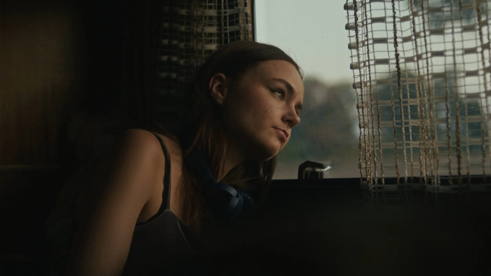 A young woman with brown hair and green eyes leaning her head against a window with curly curtains, looking outside pensively.