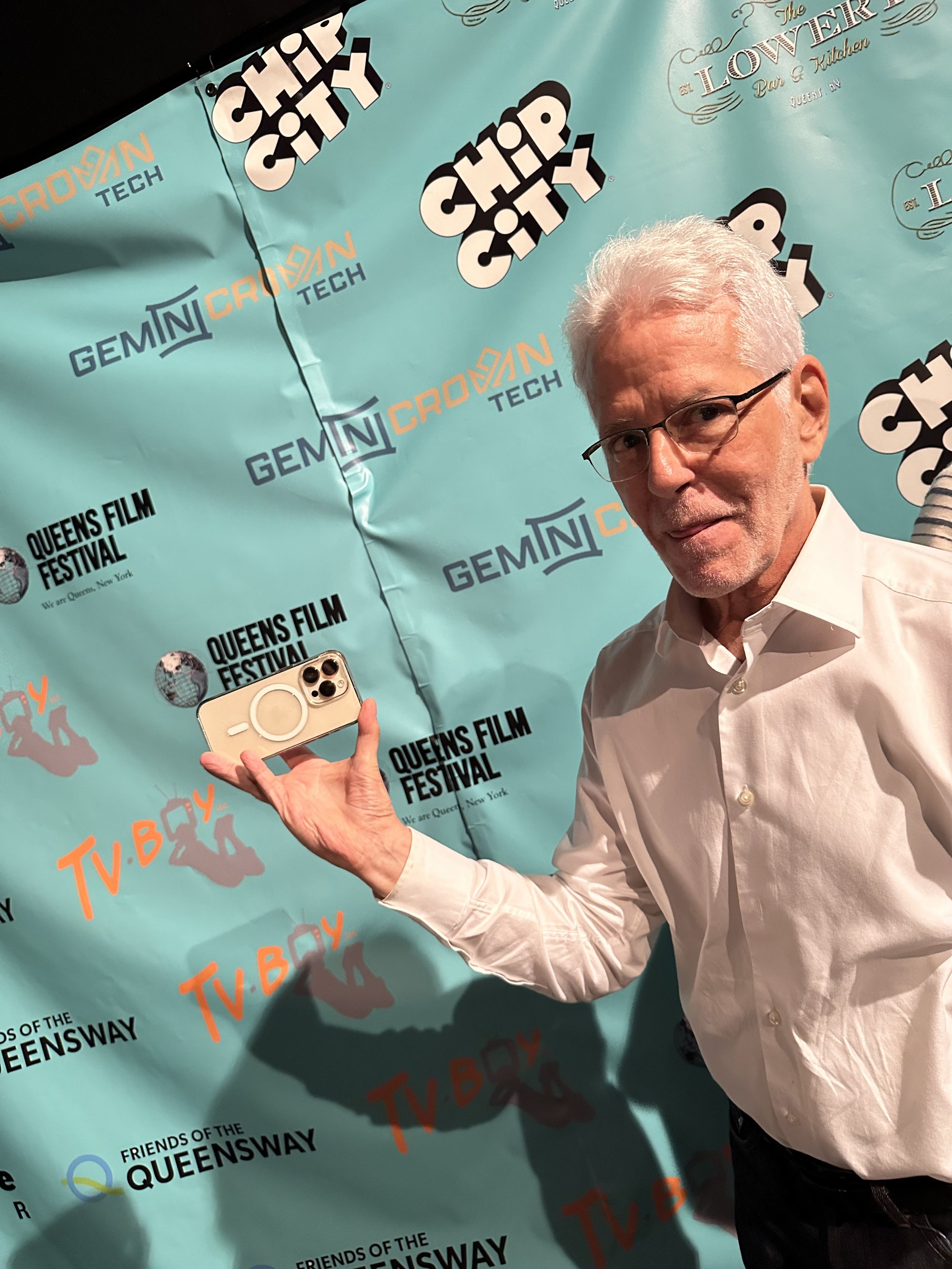 An older man with white hair and glasses holding a smartphone with a PopSocket, standing in front of a teal backdrop with event logos including 'Queens Film Festival', 'Gemini CNCrowd', and 'TV BAY'.