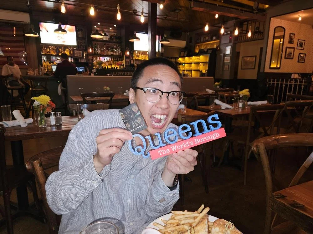 A man with glasses and a striped shirt sitting at a restaurant table, holding a Queens sign and a photo, with a plate of French fries and fried food in front of him, smiling excitedly.
