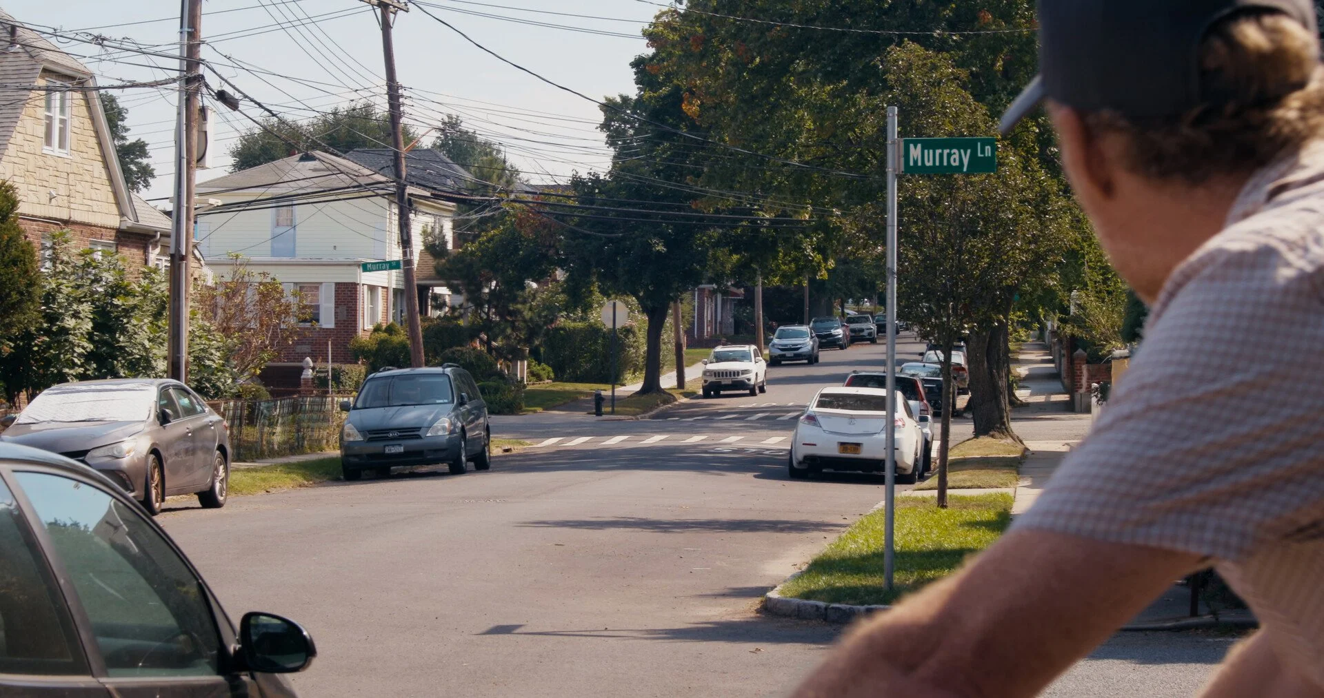 A street scene with cars parked on both sides of the street, trees, houses, and a person in the foreground wearing a cap and leaning over, with a sign indicating the intersection at Murray Lane.